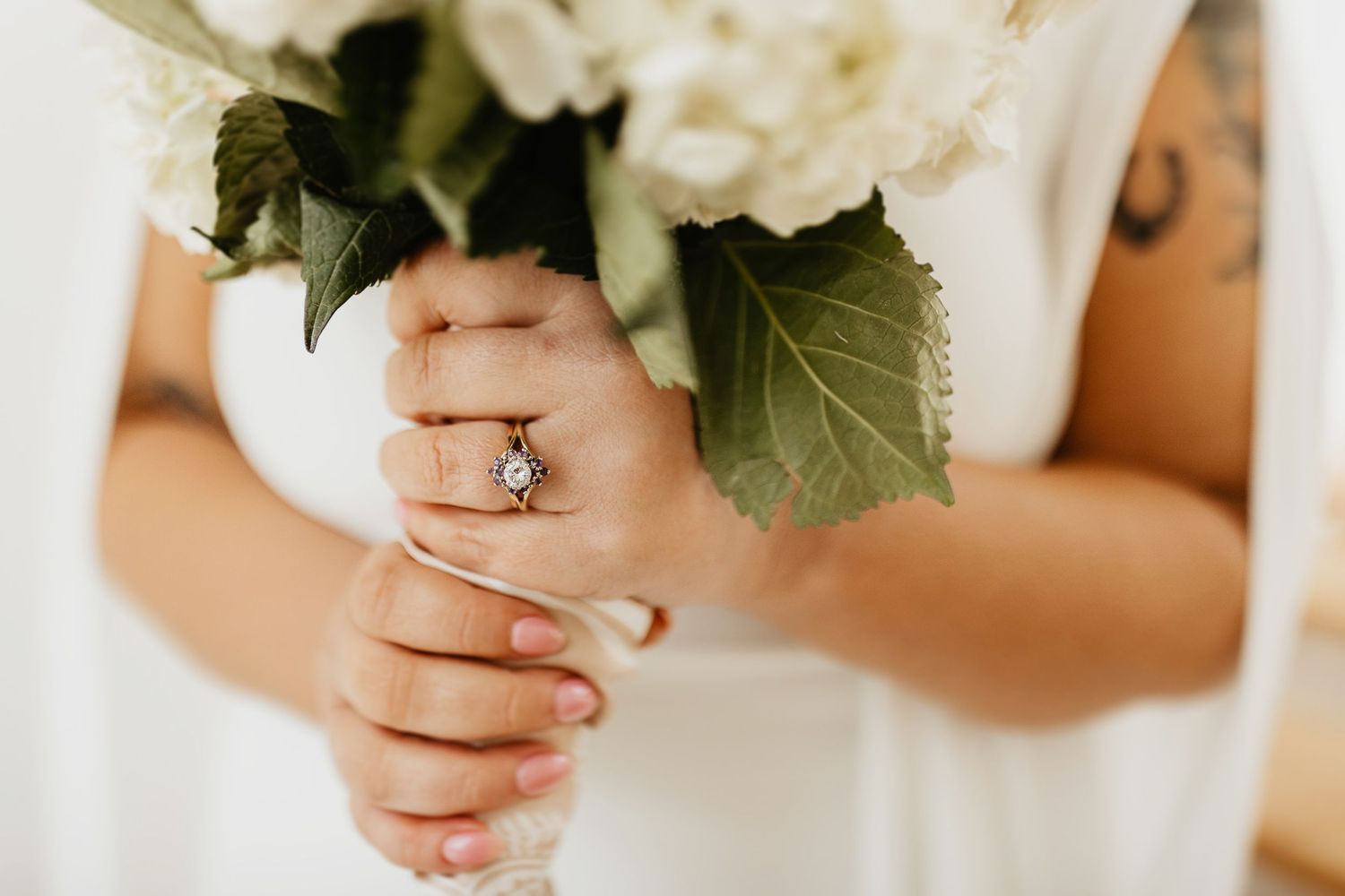 Close-up view of hands holding a white wedding bouquet with green leaves, displaying an elegant wedding ring.