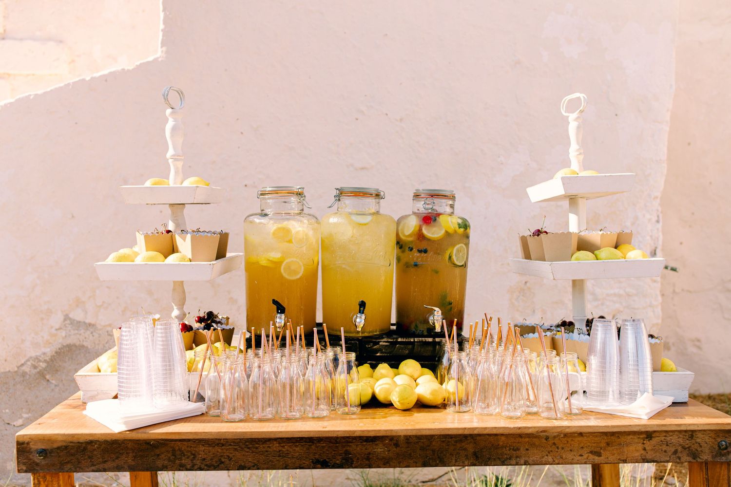 Rustic wooden table set with glass drink dispensers, white tiered dessert stands, and cocktail glasses for a party setup.