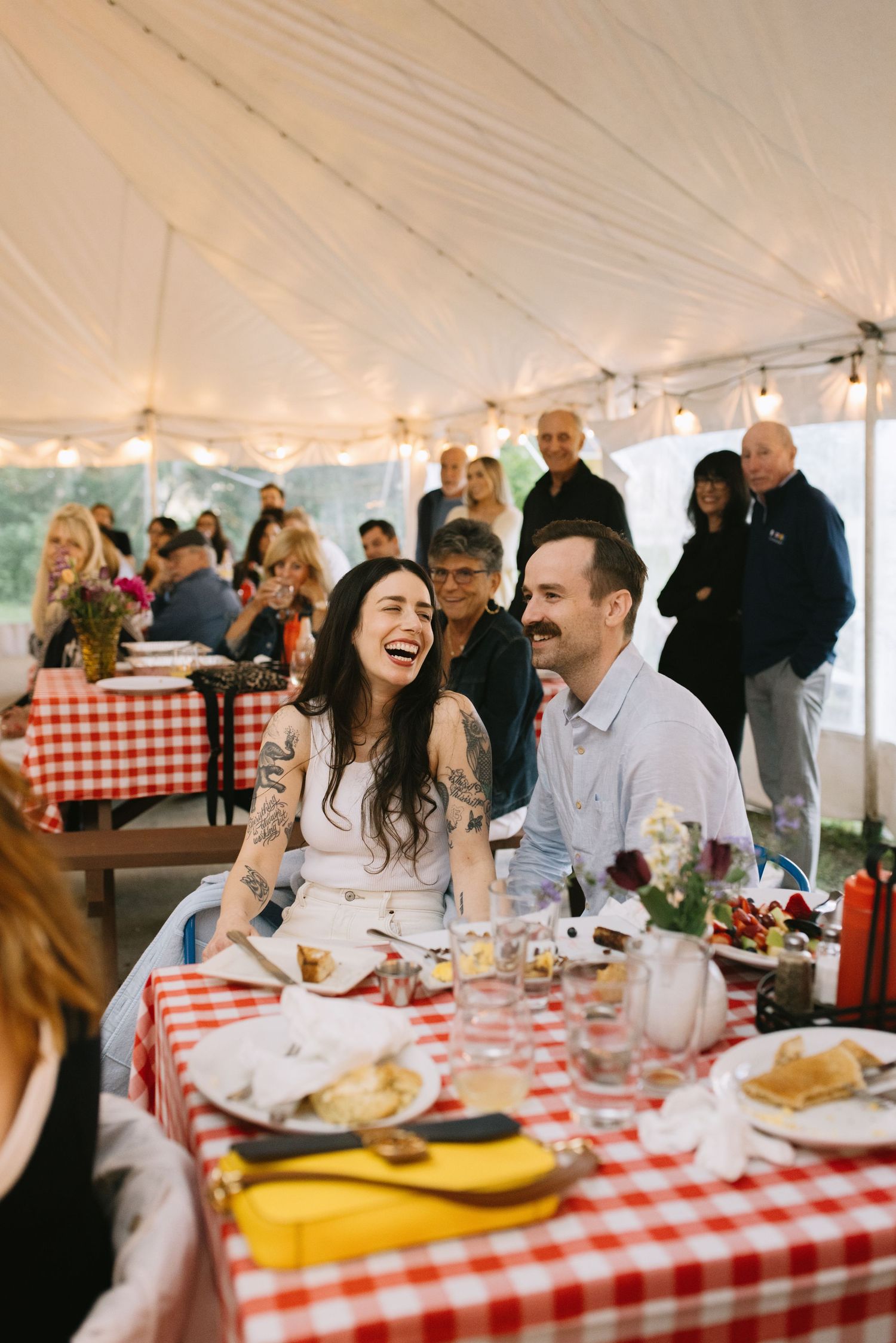 People enjoying a casual outdoor dinner under a white tent with red checkered tablecloths and rustic decor.