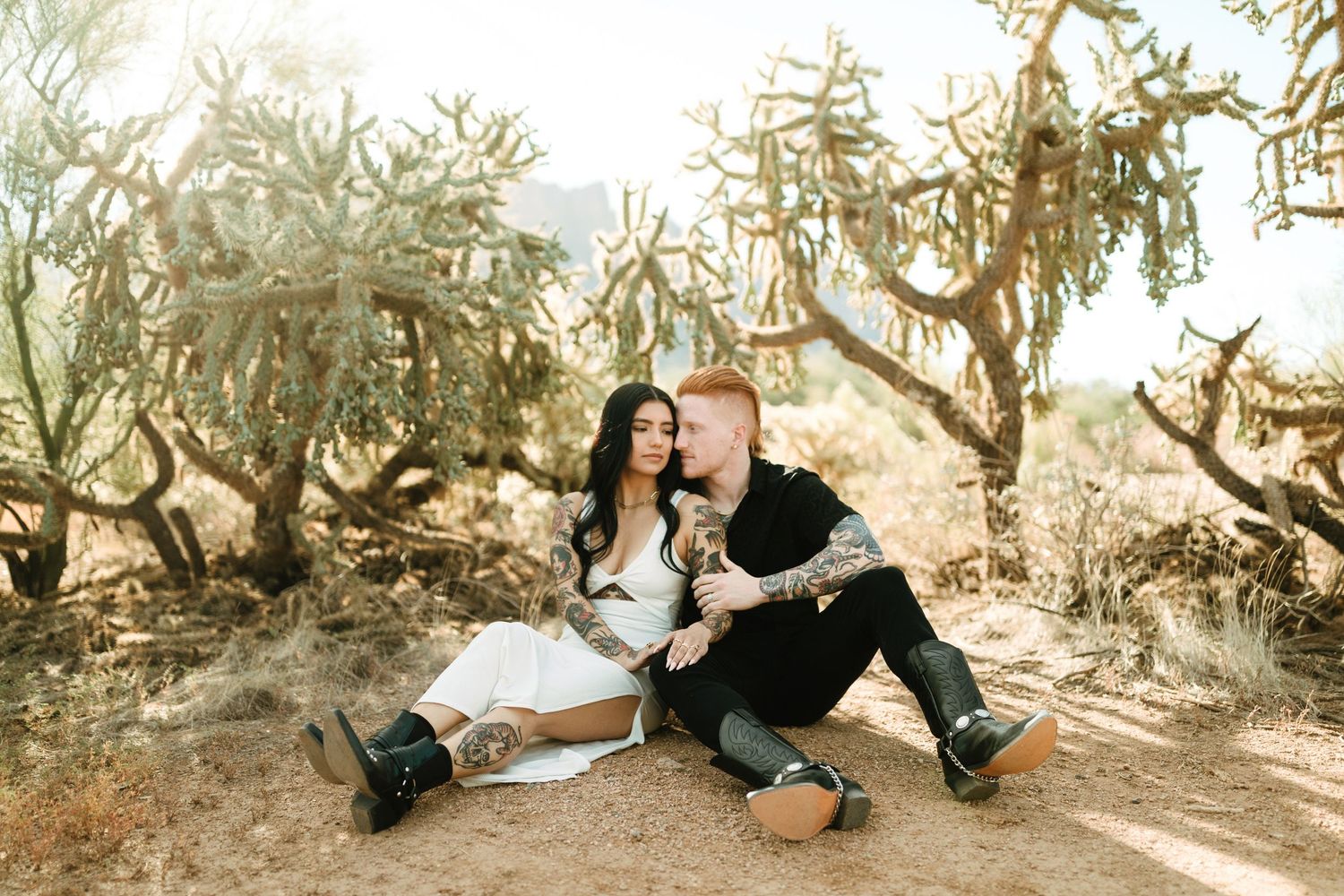 A couple sits intimately together on the ground amongst a grove of dried winter trees.