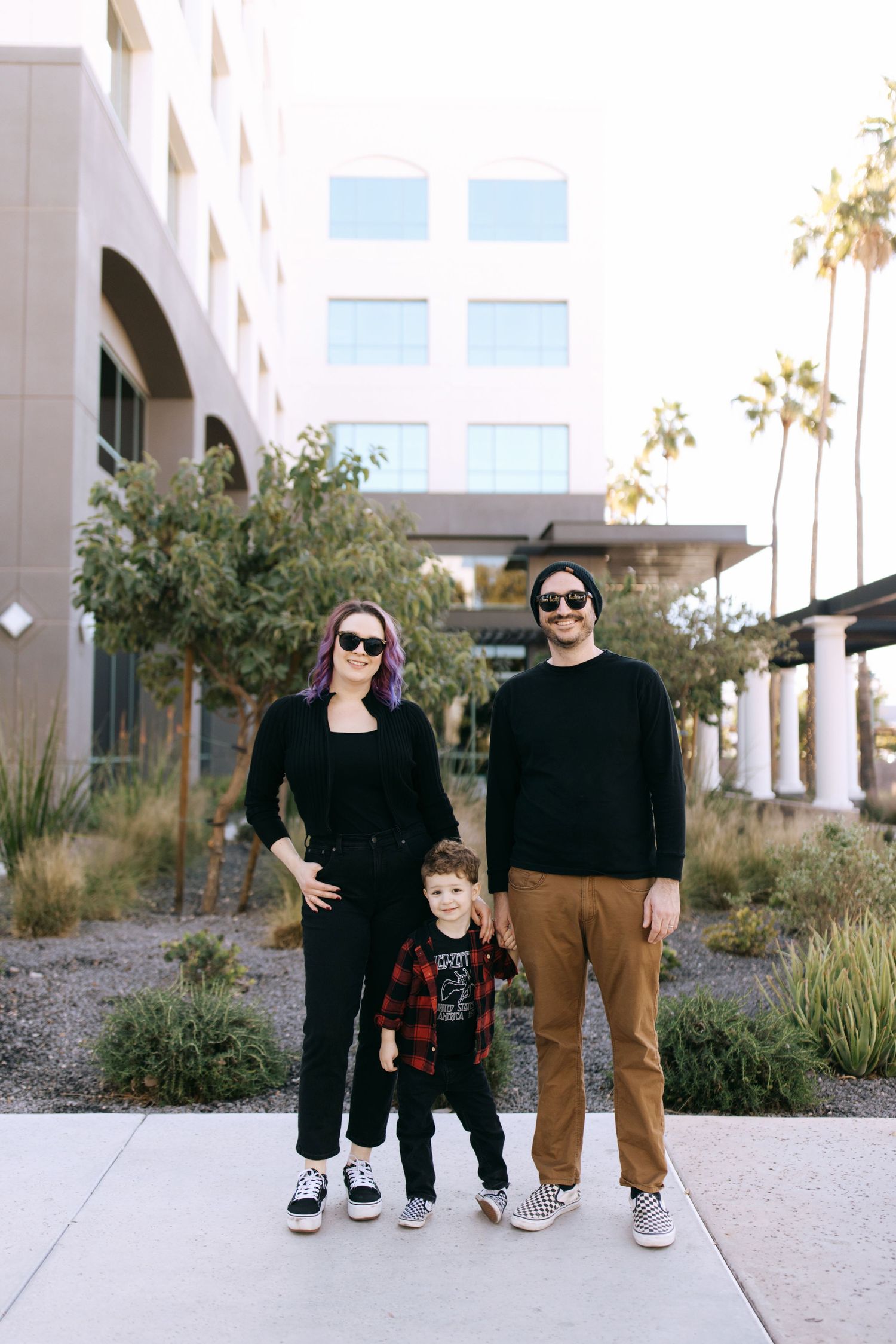 A family of three wearing black tops stands together outside a modern building with palm trees in the background.