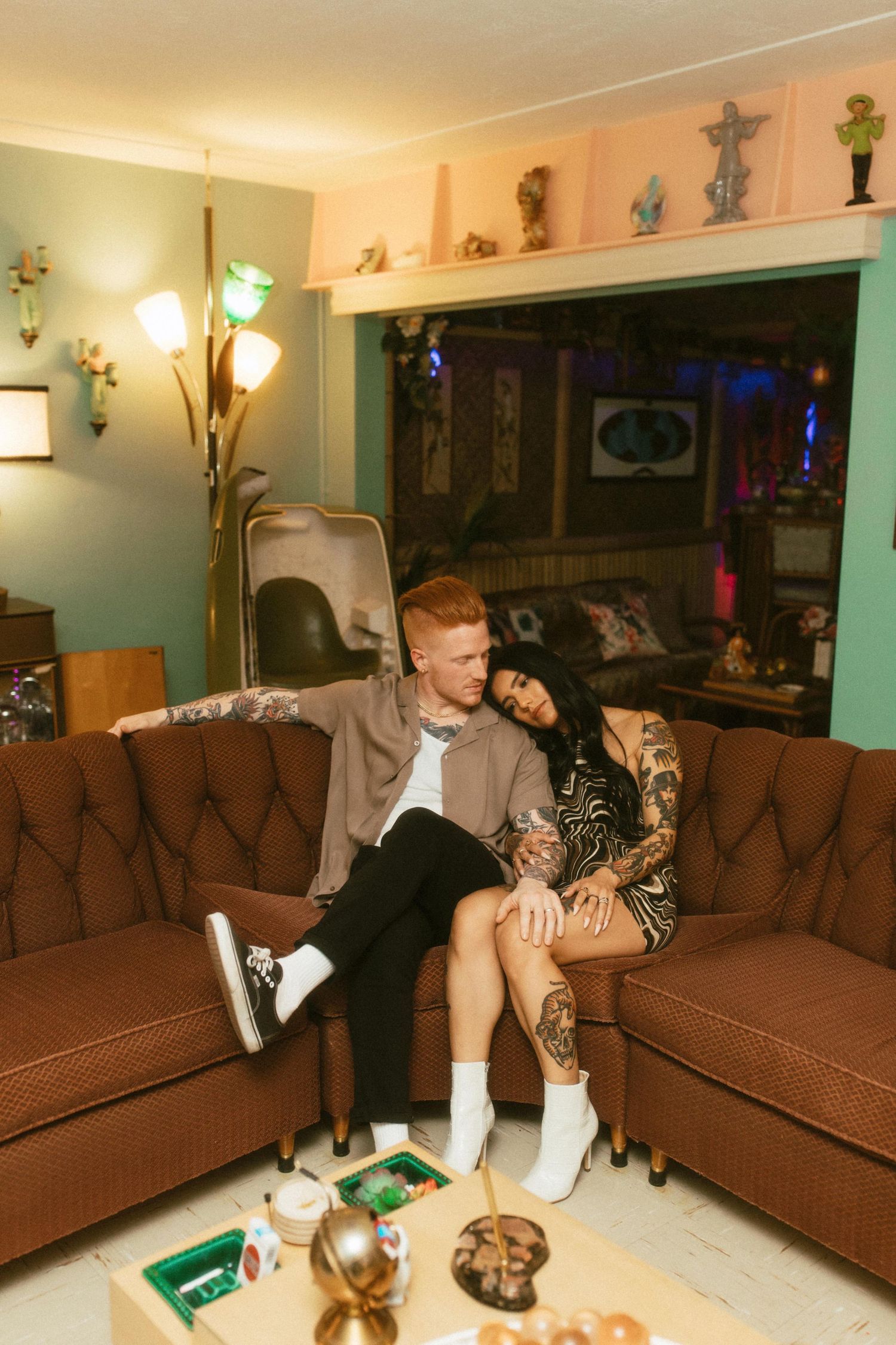 A couple relaxes together on a brown couch in a dimly lit living room with decorative figurines displayed on wall shelves.