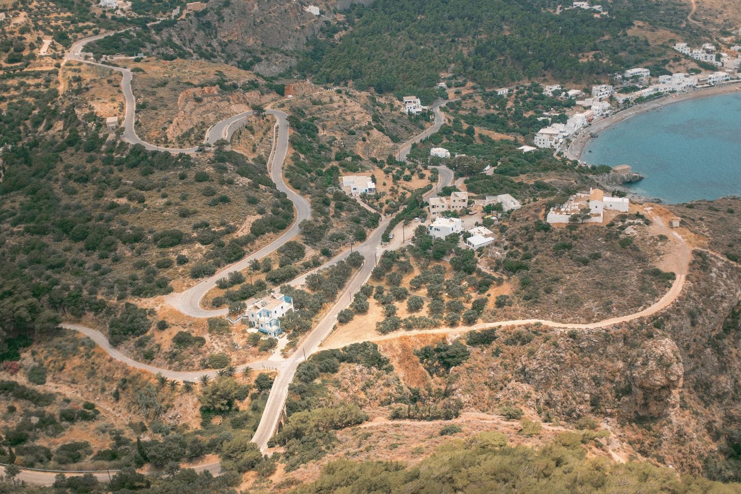 Aerial view of winding coastal roads and buildings along a Mediterranean hillside with turquoise waters in the distance.