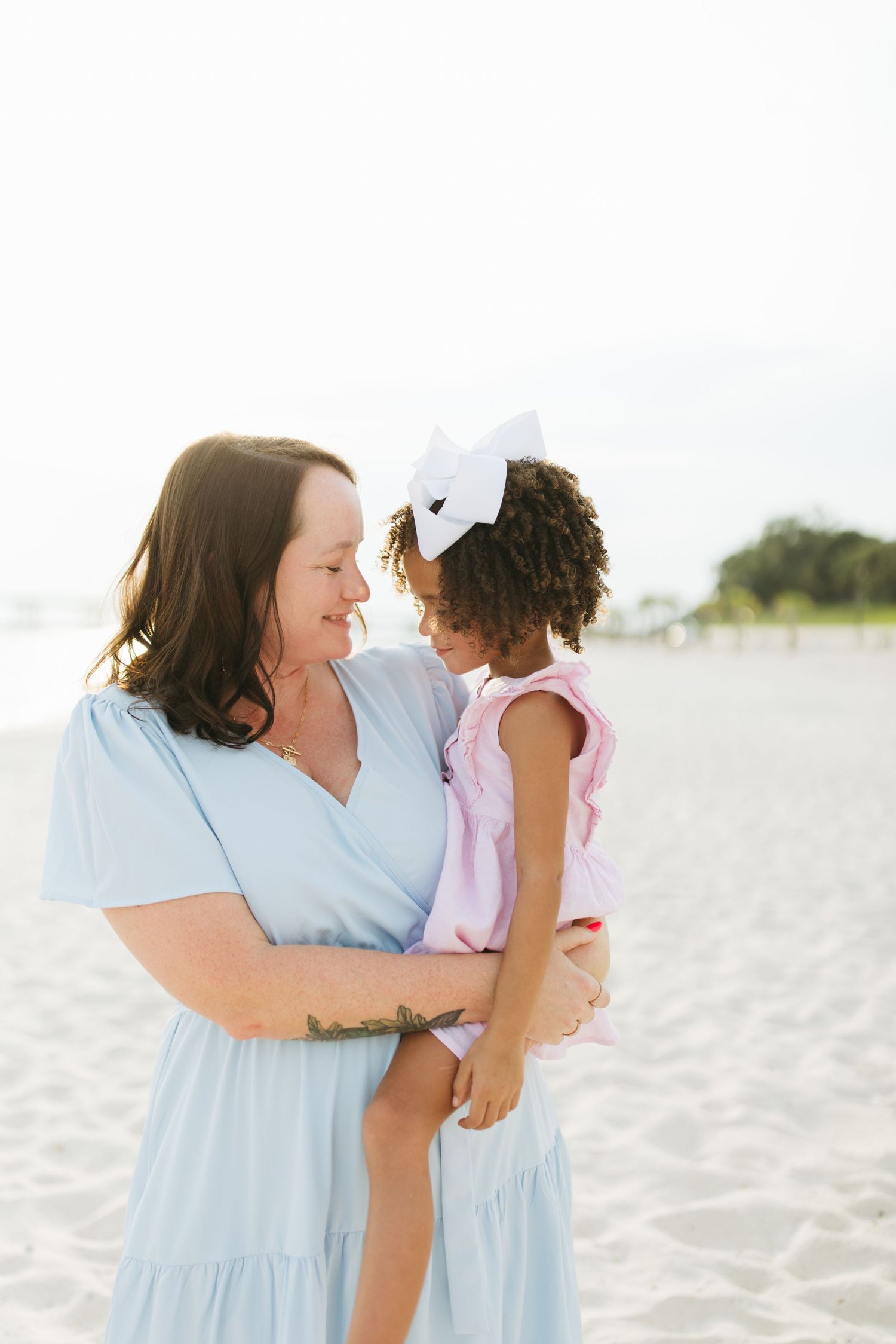 Mother and child share a tender moment together while standing on a sunny beach in light blue and pink outfits.