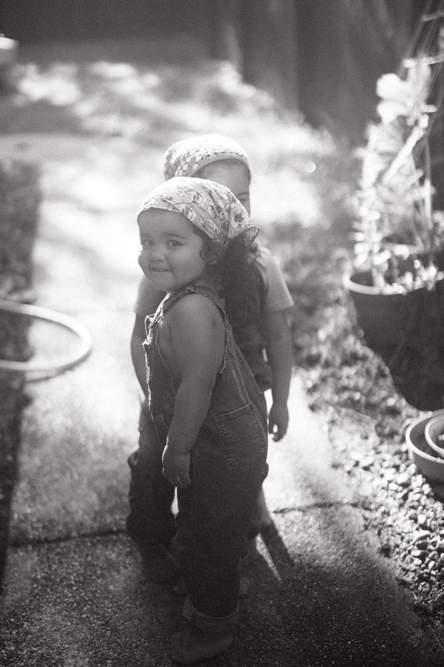 A black and white photo of a child in overalls and a headscarf standing in dappled sunlight outdoors.