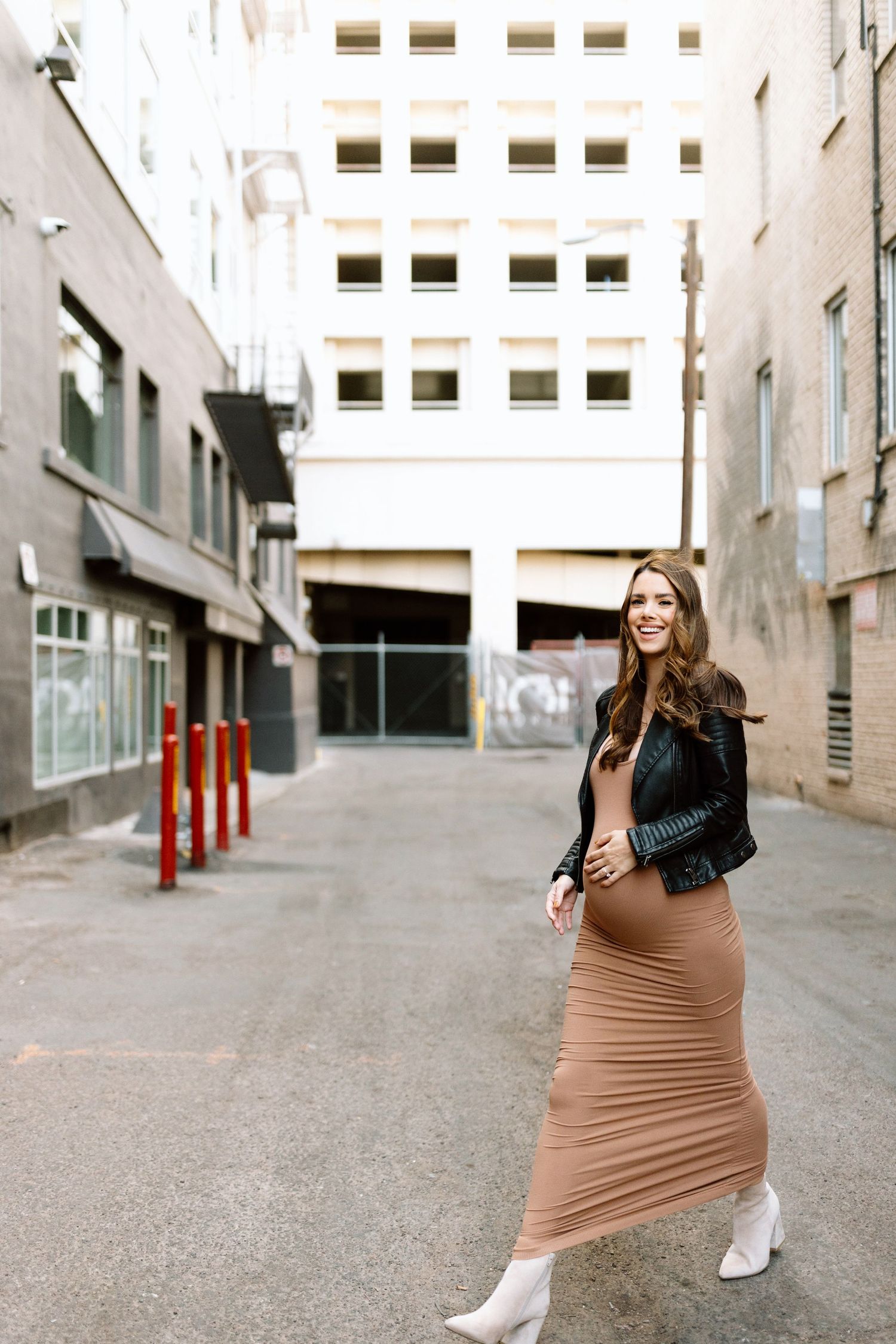 Fashionable woman in a form-fitting tan dress and black jacket poses on an urban street with tall buildings.