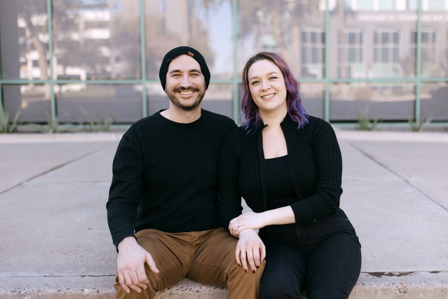 A couple sitting together outdoors on a concrete surface, with the person on the right having vibrant purple hair.