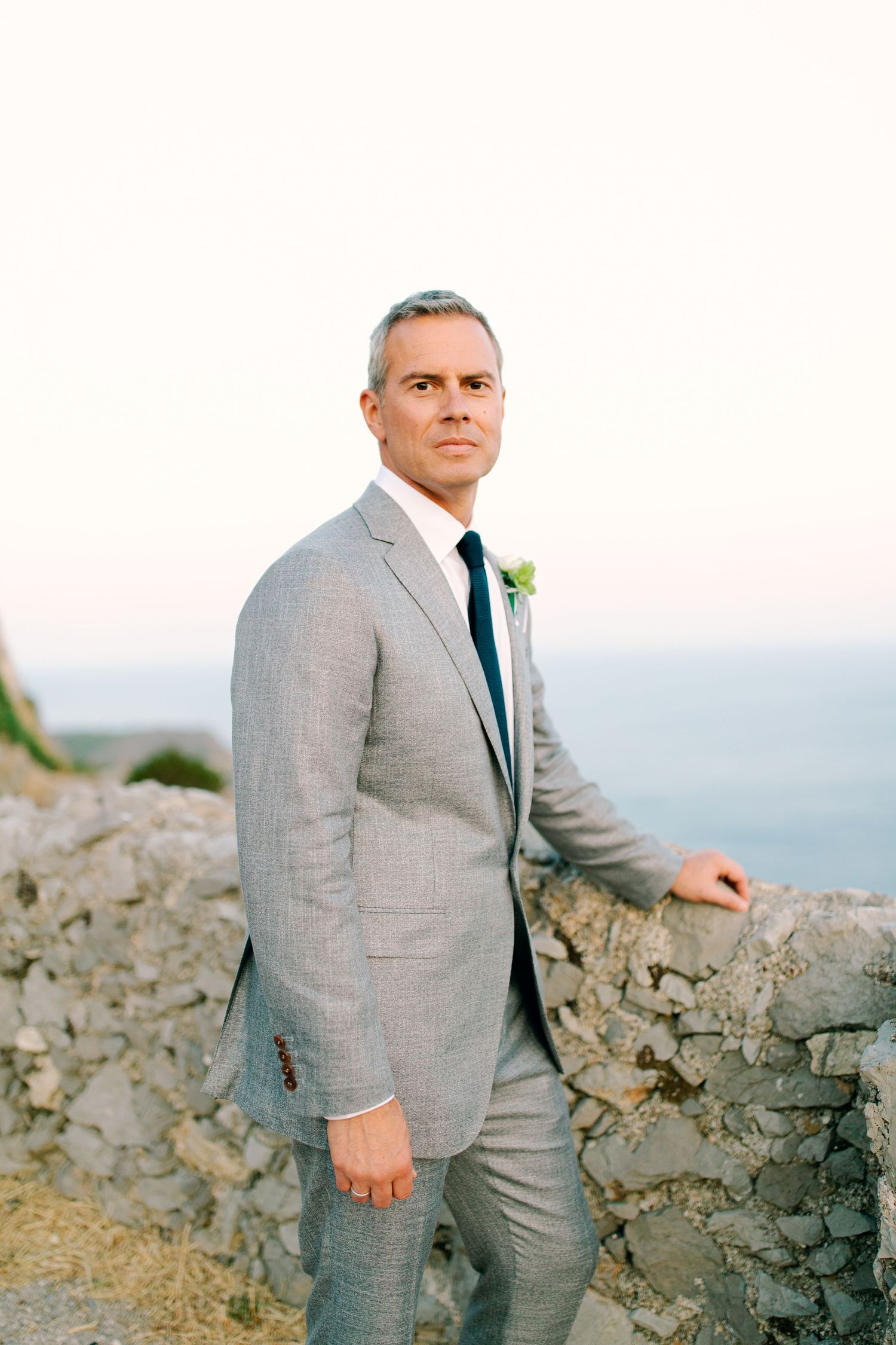 A groom in a light gray suit with a boutonniere poses against a rocky coastal backdrop at sunset.