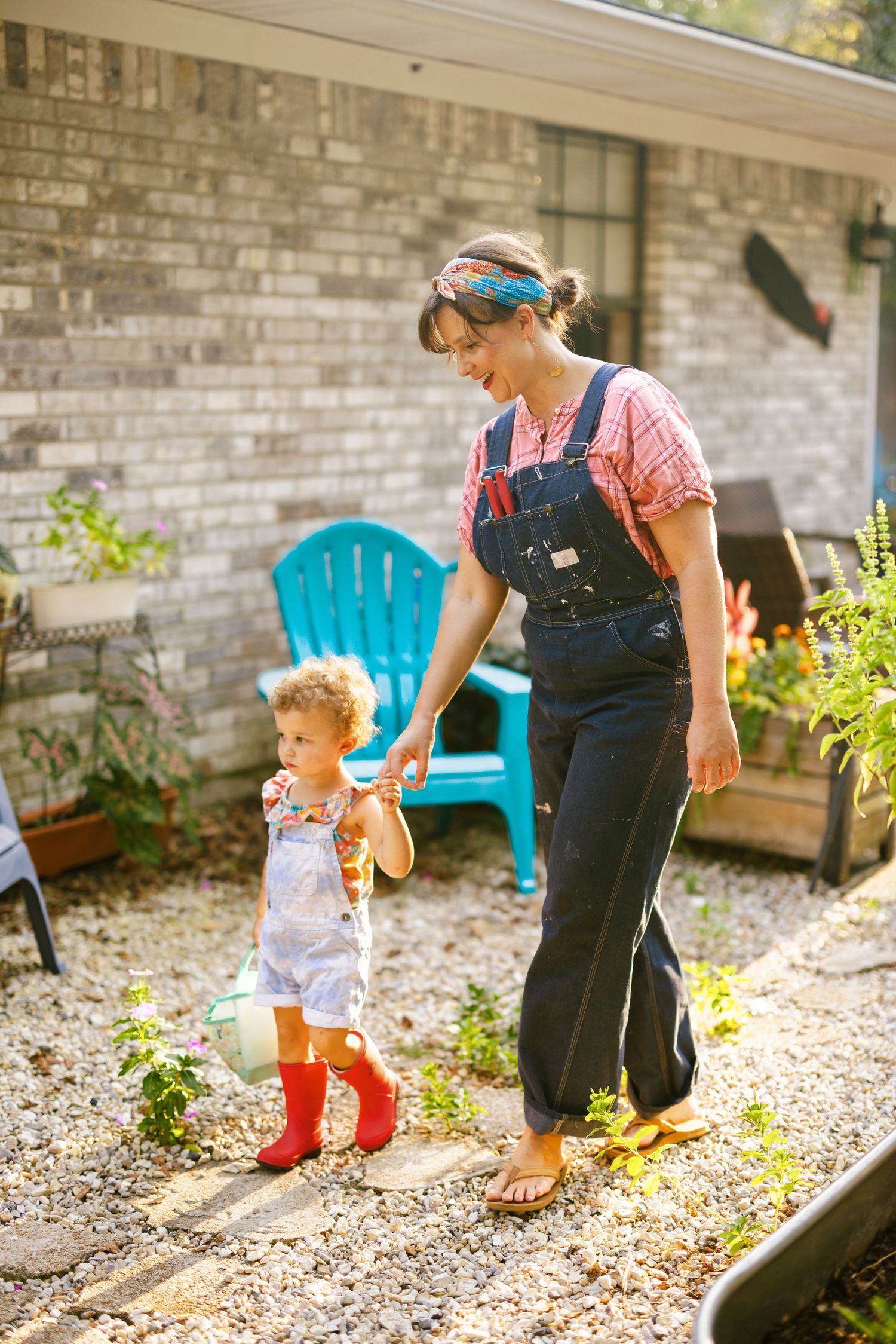 Two people explore a garden bed outside a brick house with a bright turquoise chair visible in the background.