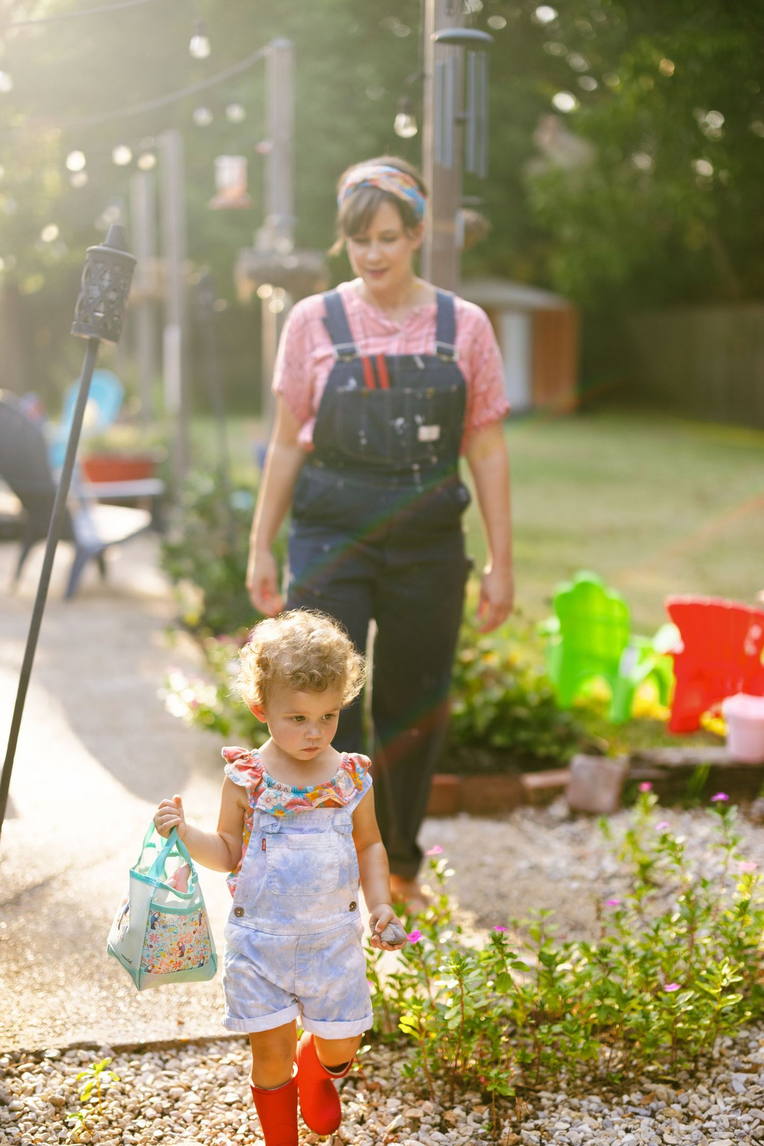 A young child in a floral outfit plays with watering cans and toys in a garden while being supervised.