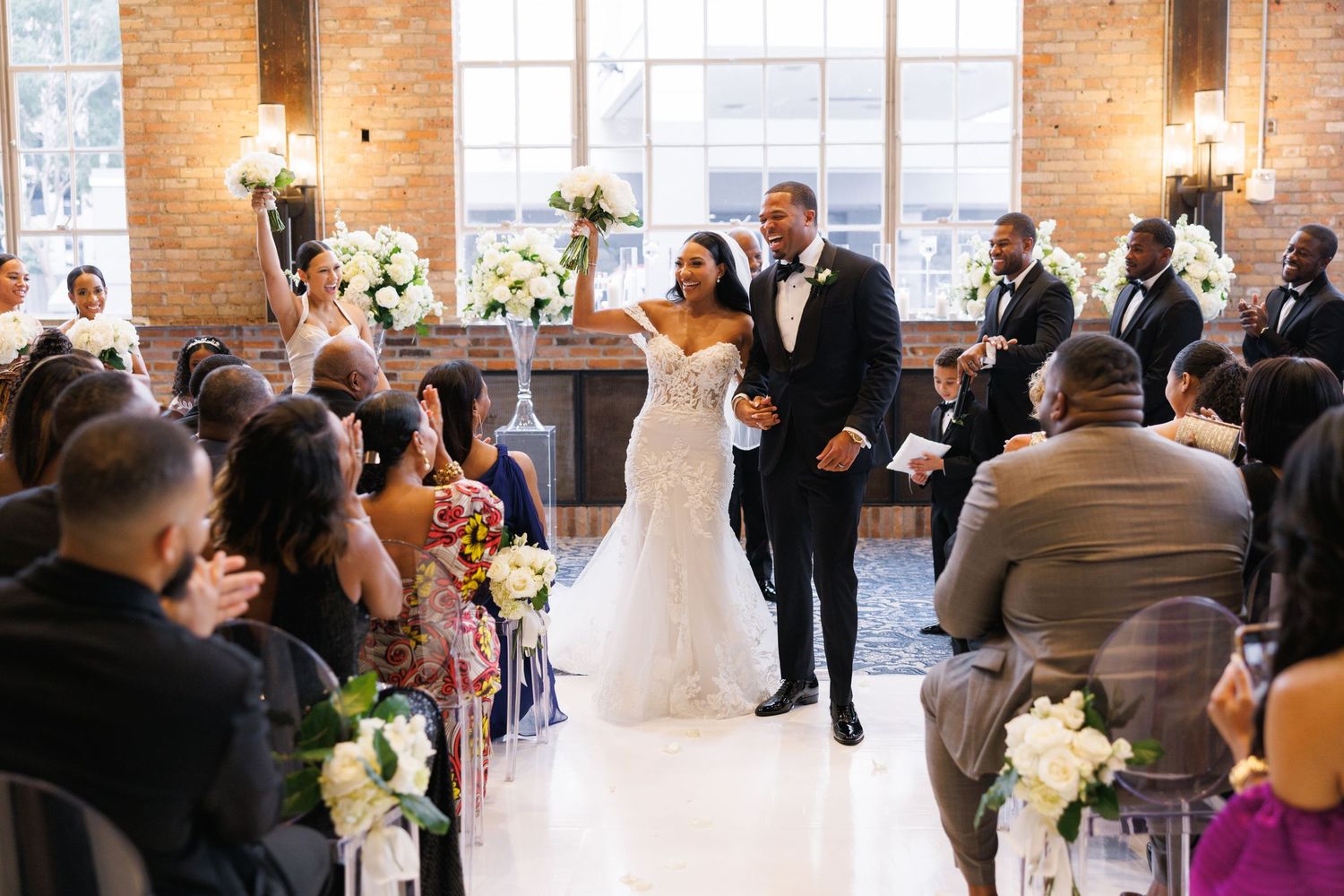 Elegant wedding ceremony takes place in industrial brick venue with floral arrangements and white aisle runner.