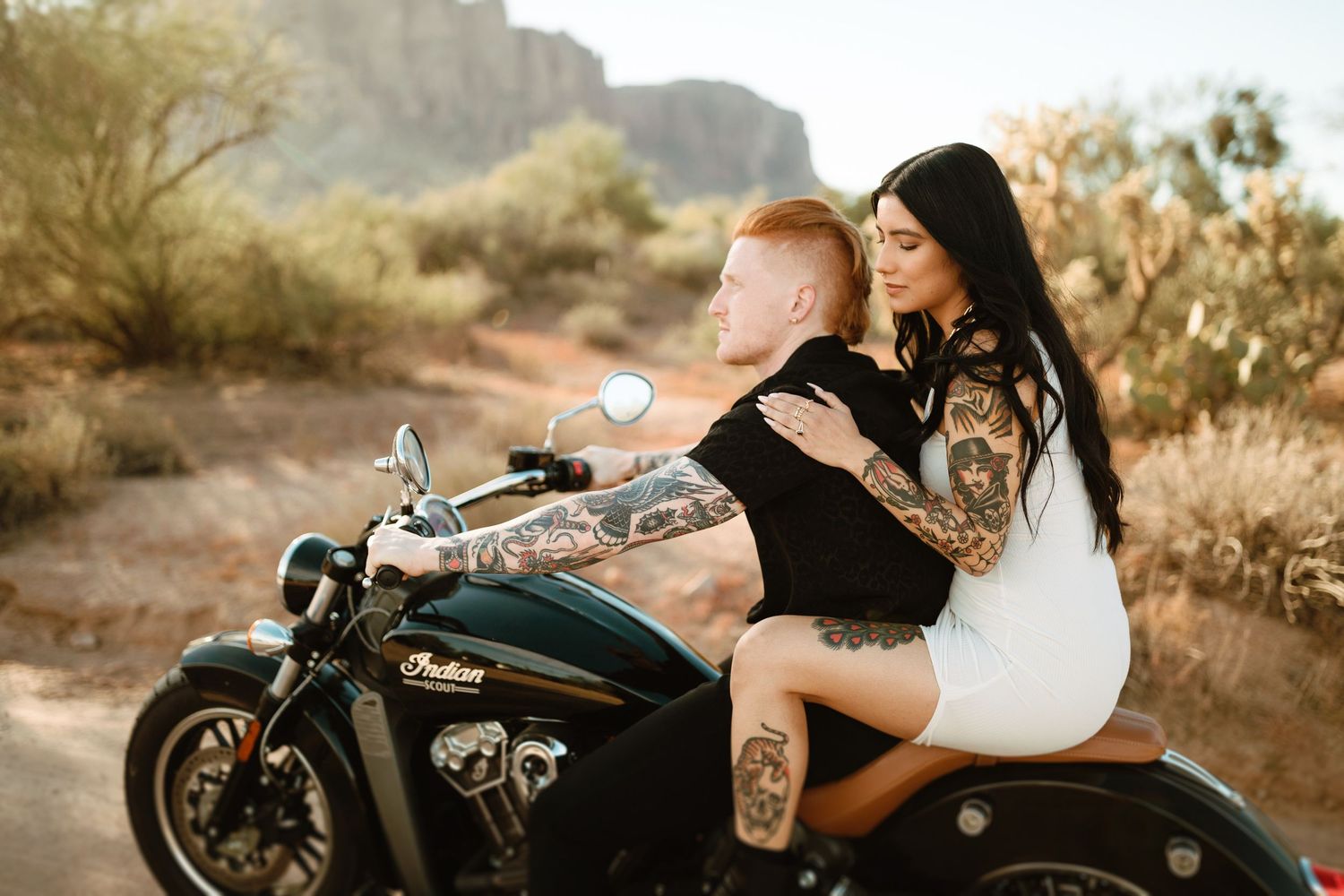 A couple rides together on an Indian motorcycle in a desert landscape with mountains in the background at sunset.
