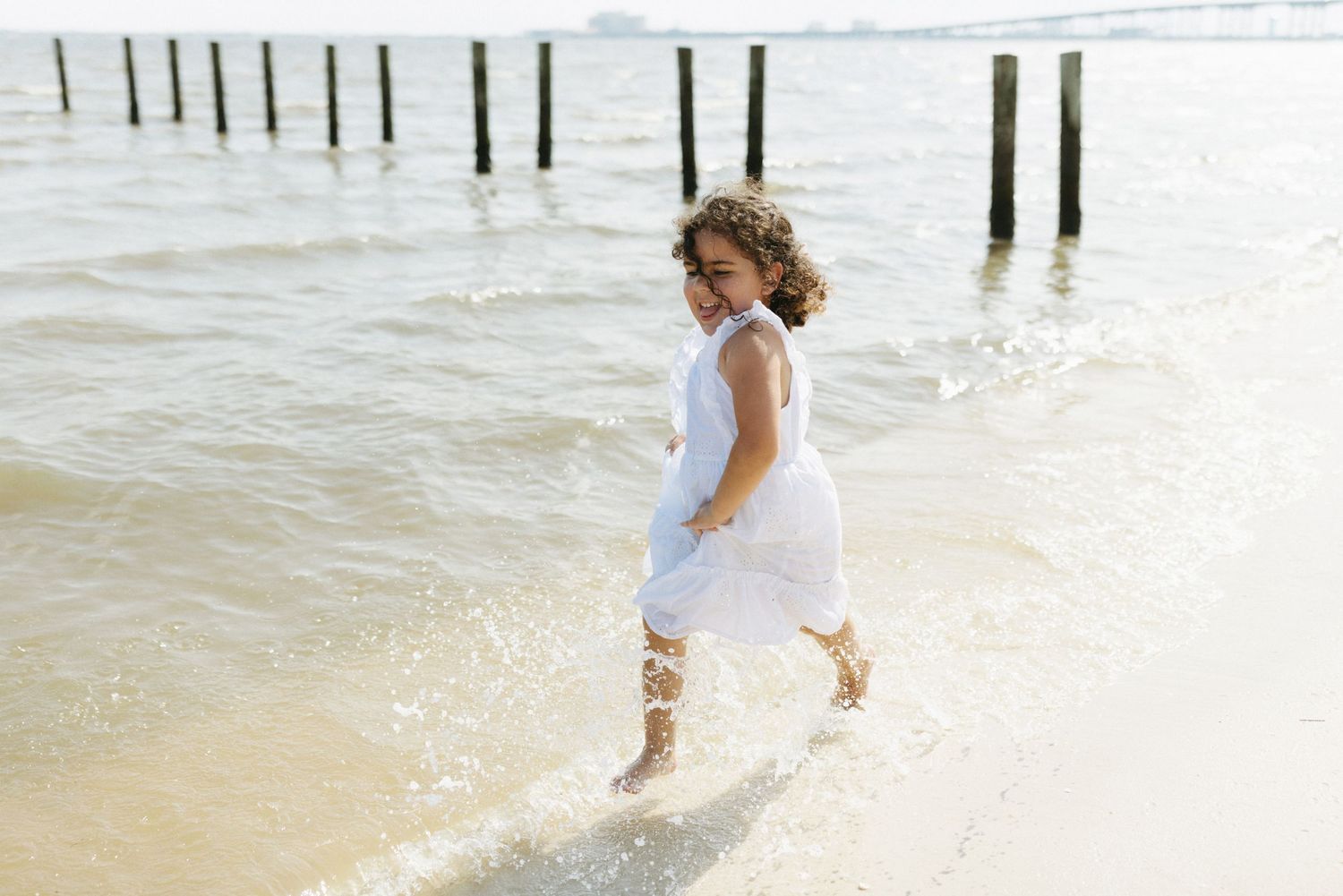 A person in a white dress splashes joyfully through shallow ocean water near wooden pier posts.