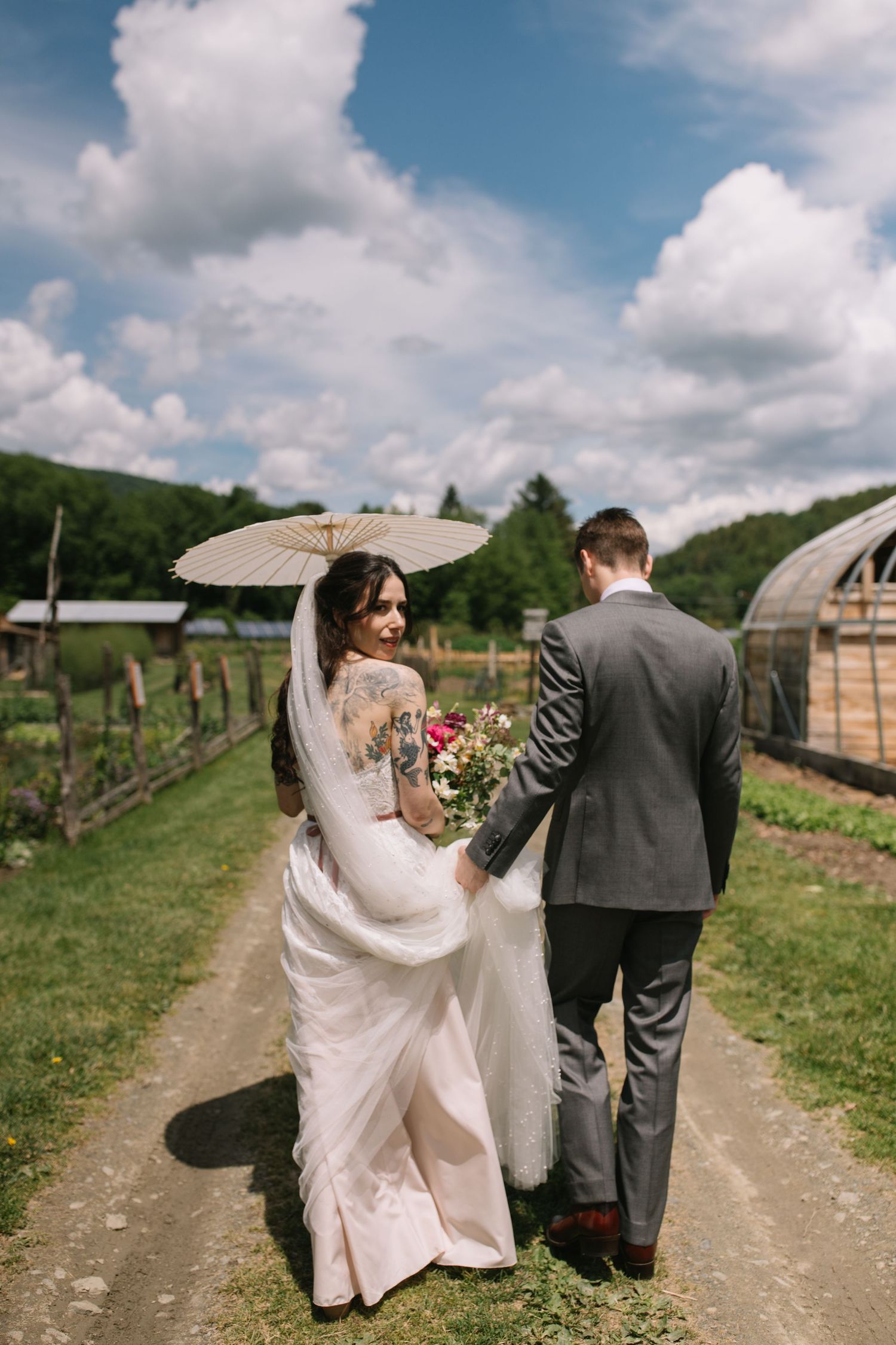 A couple on their wedding day walks down a dirt path at a rustic farm with greenhouses and mountains in the background.