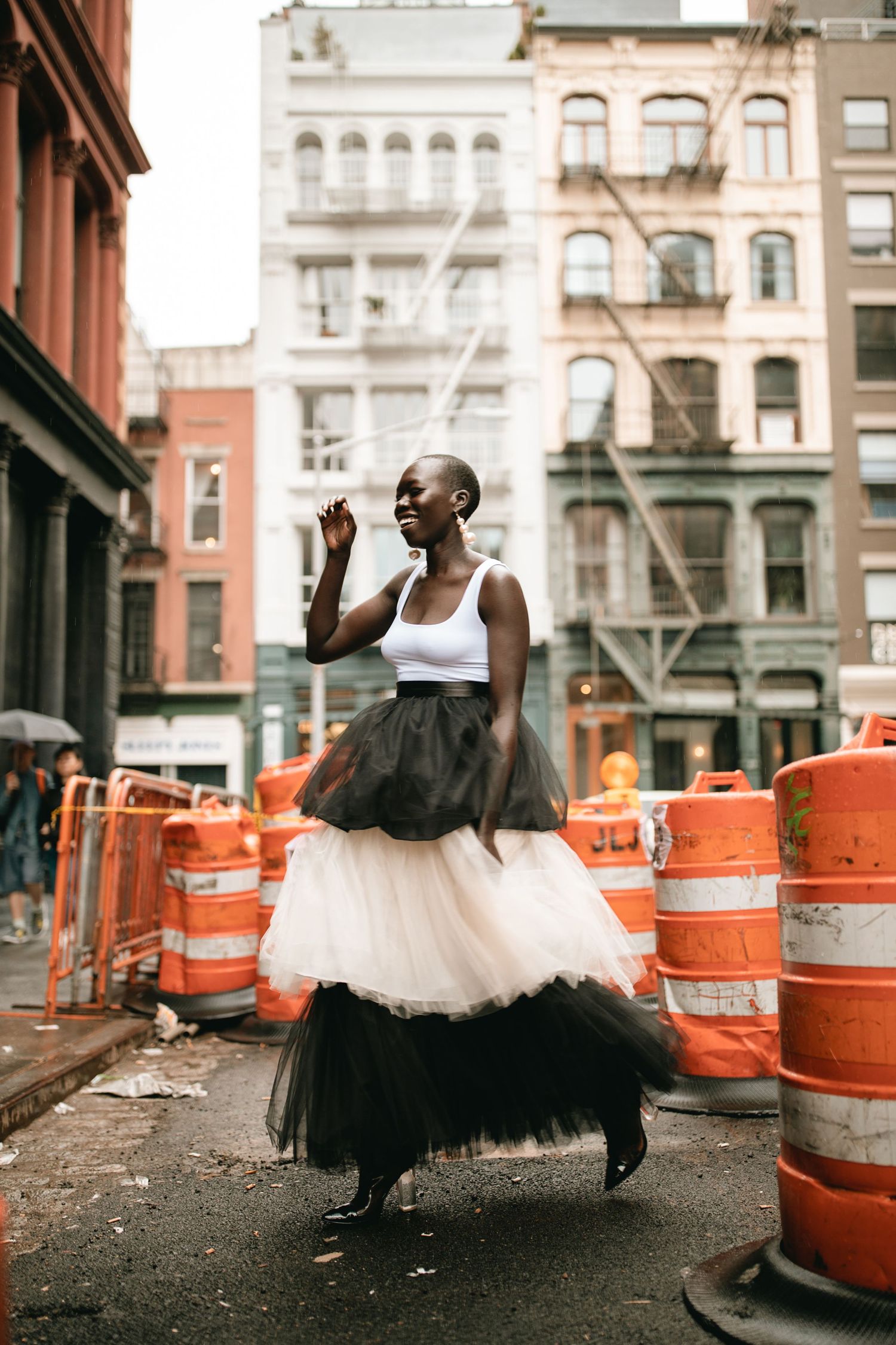 A dancer in a black and white tulle dress poses gracefully amid orange construction barriers on a New York City street.