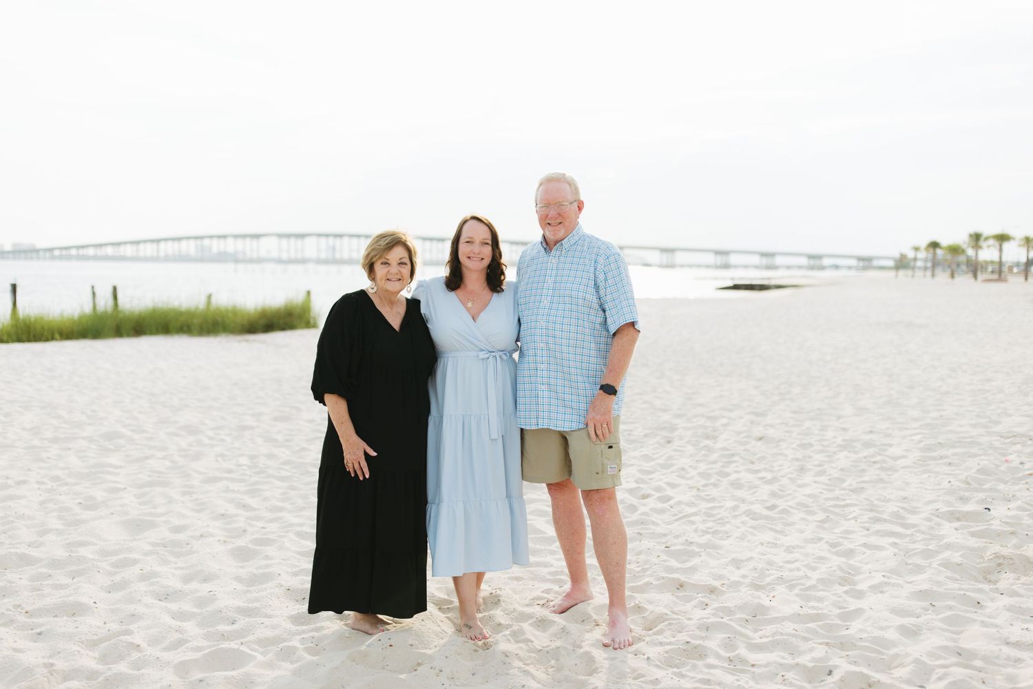 Three people standing together on a white sandy beach with palm trees and a pier visible in the background.
