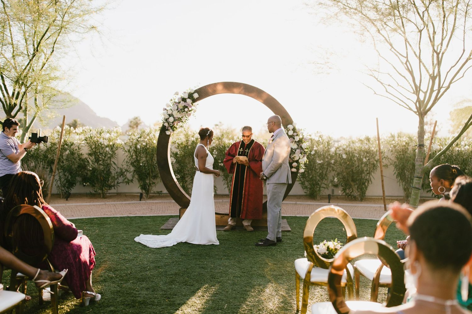 Outdoor wedding ceremony under string lights as sun sets behind an elegant floral arch in a garden setting.