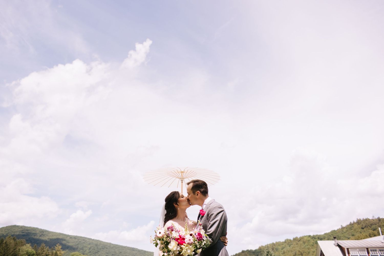 A couple shares a romantic kiss on a mountaintop surrounded by scenic mountain vistas and dramatic cloudy skies.