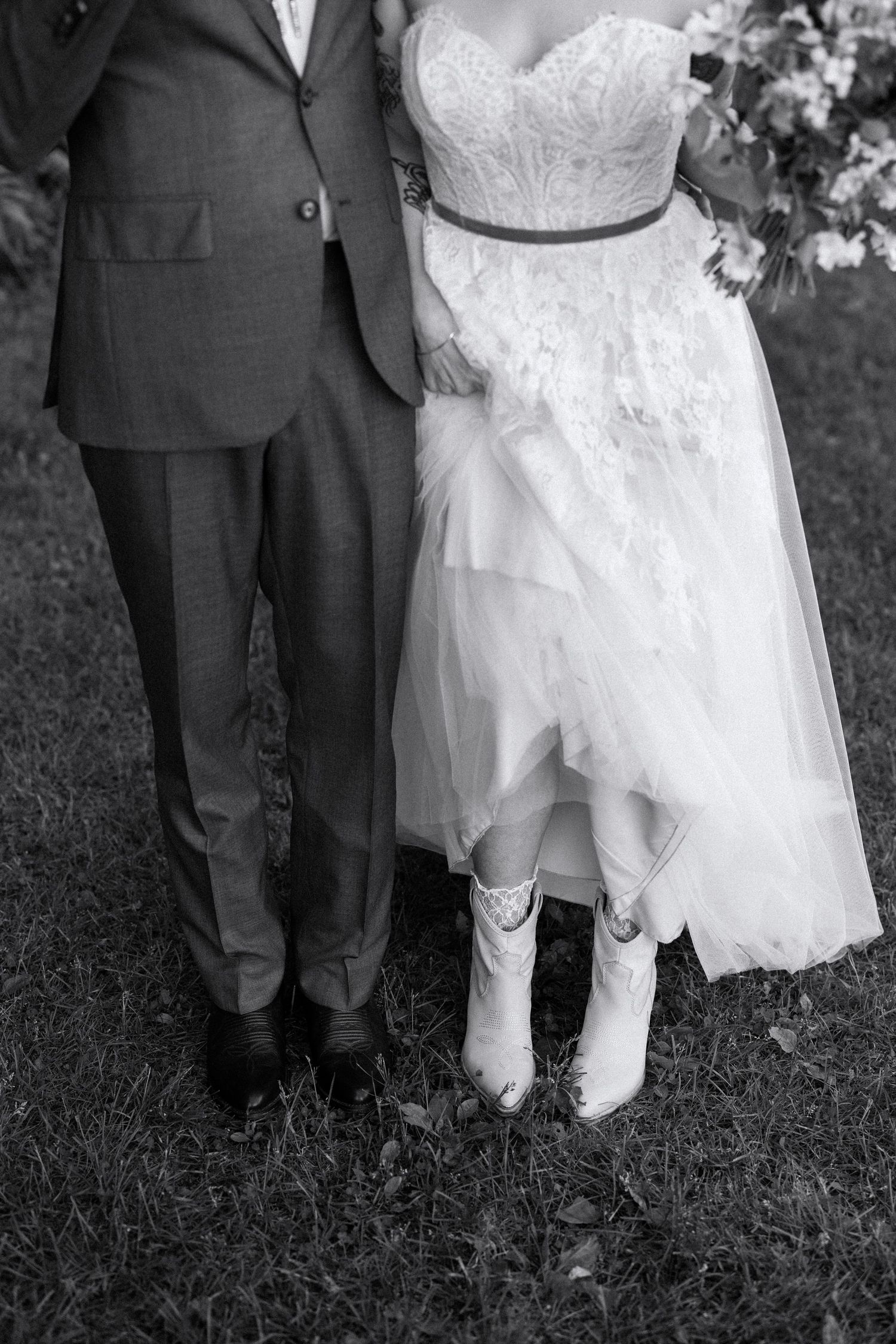 Black and white wedding photo showing bride in cowboy boots with lace dress next to groom in formal suit standing on grass.