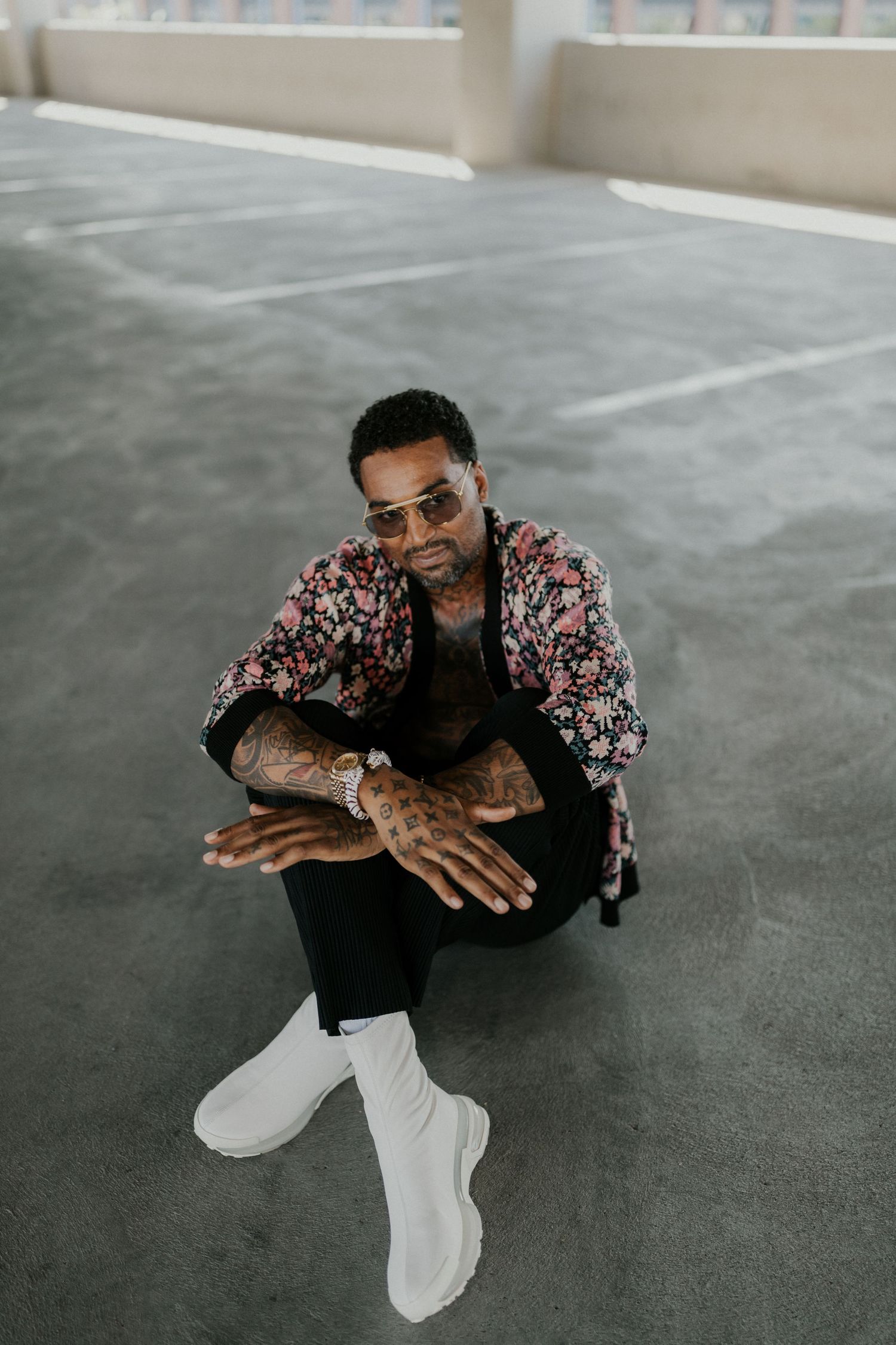 A person wearing a floral print shirt and white pants poses expressively on a gray concrete floor.