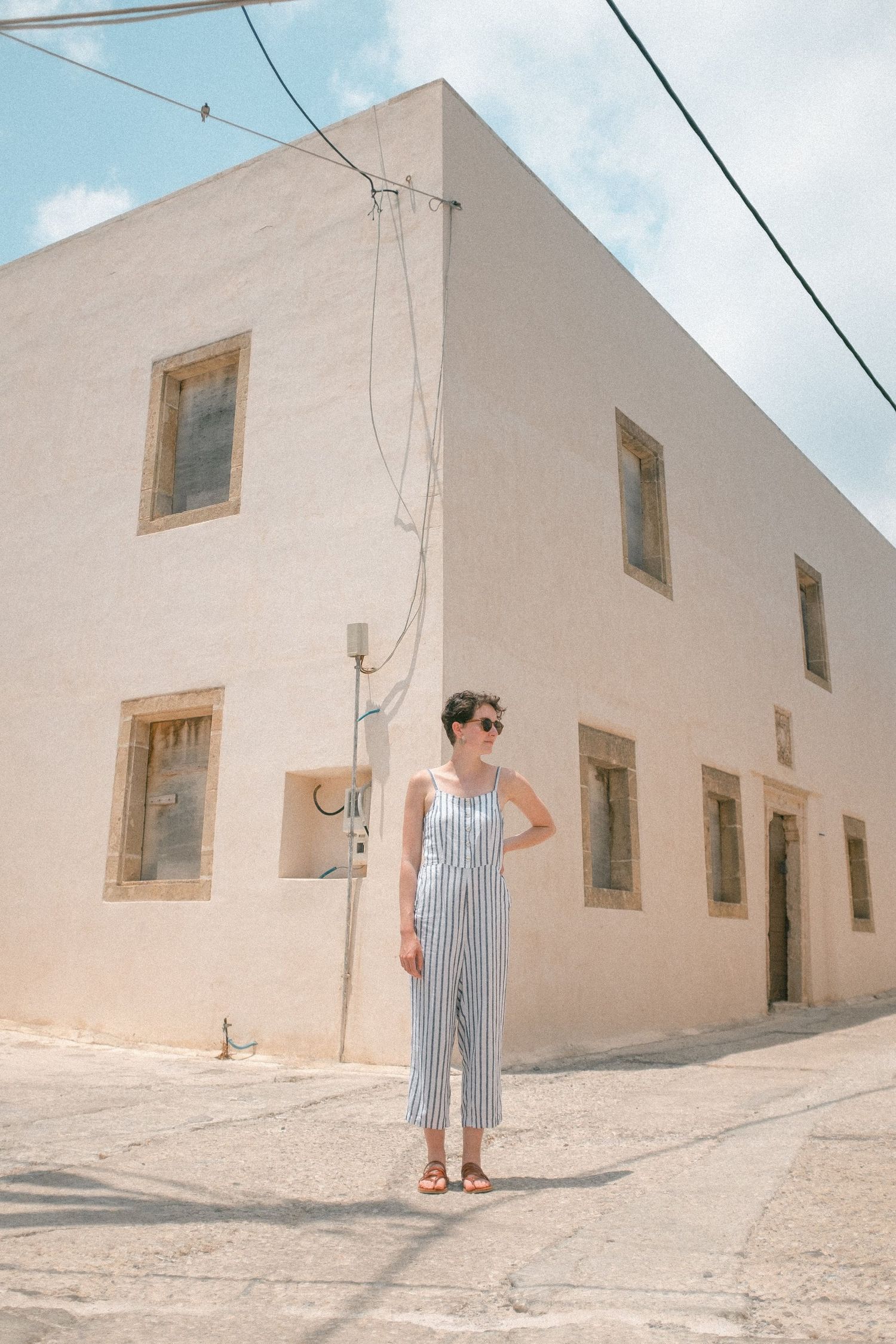 A person in striped jumpsuit stands on a corner by a whitewashed Mediterranean building on a sunny day.