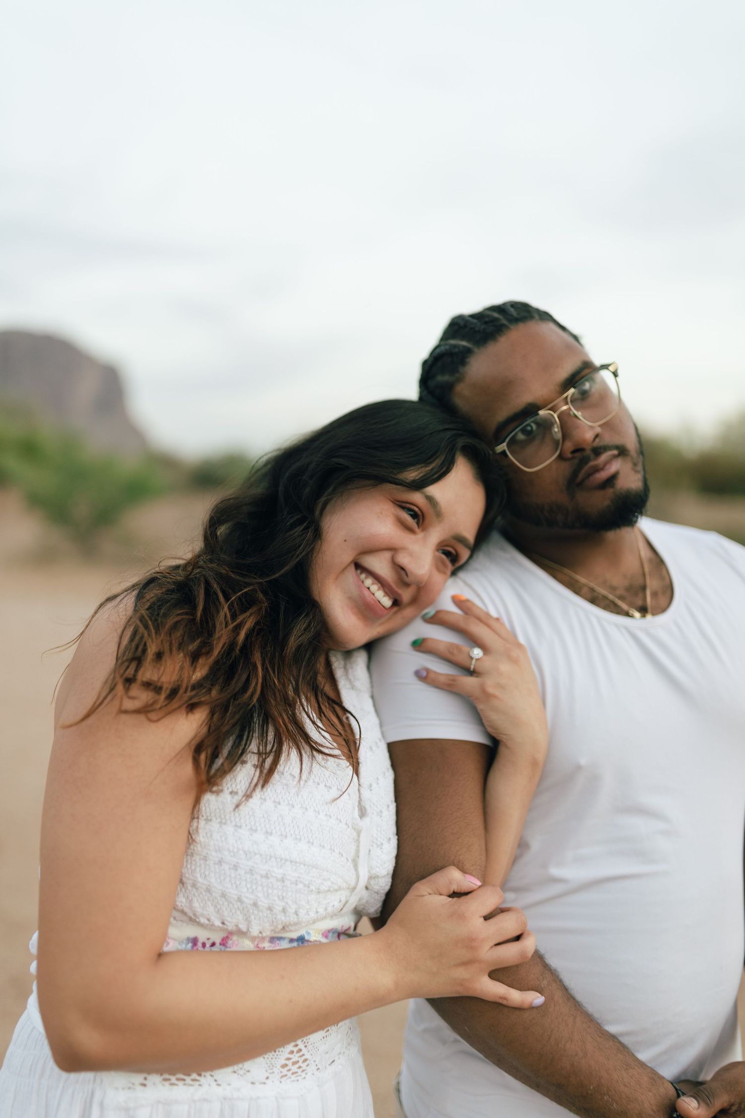A happy couple embraces and laughs together while sitting outdoors in casual white outfits on a sunny day.