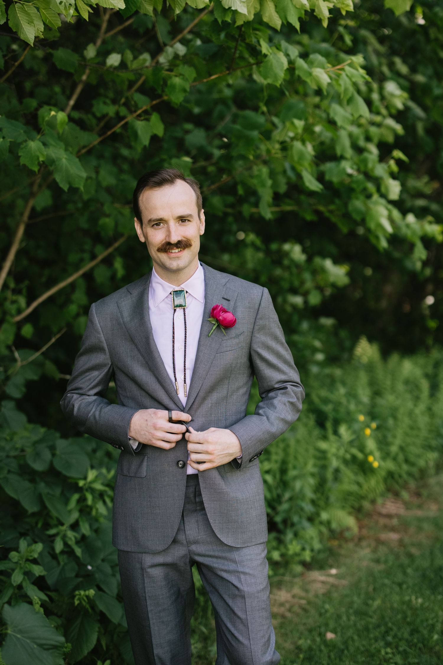 A man in a gray suit and tie poses outdoors with a pink flower bouquet in front of lush green foliage.