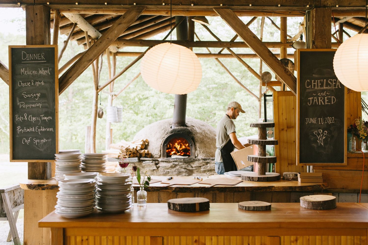 A rustic wood-fired pizza station with stacked plates and chalkboard menus in an outdoor covered dining area.