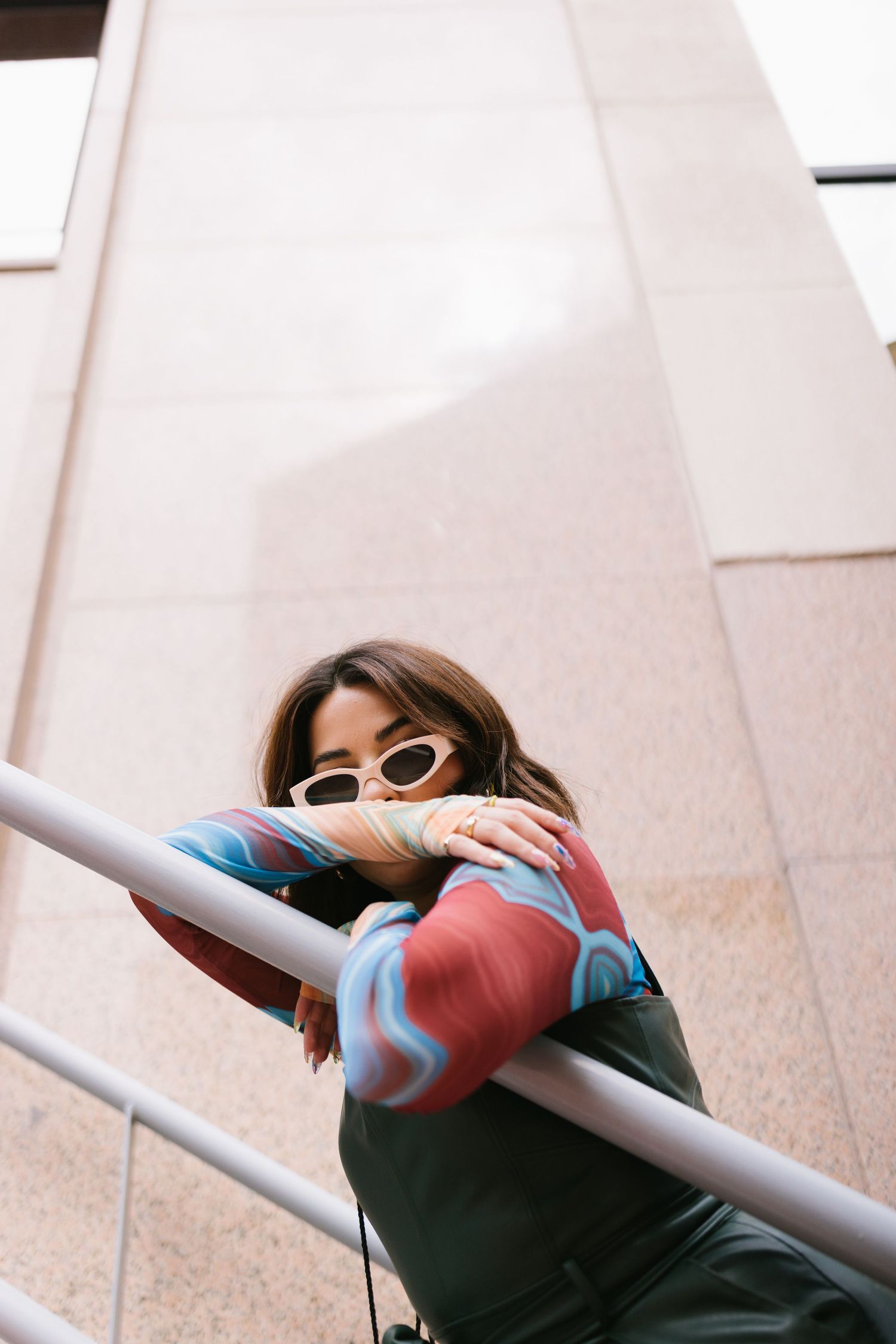 Someone wearing retro sunglasses and a colorful sweater leans against a metal railing in front of a beige building.