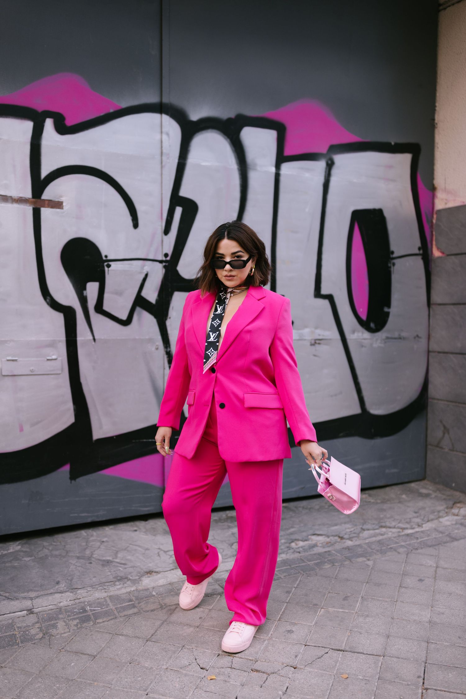 Fashionable person in vibrant hot pink suit strikes dynamic poses against graffiti wall wearing sunglasses.