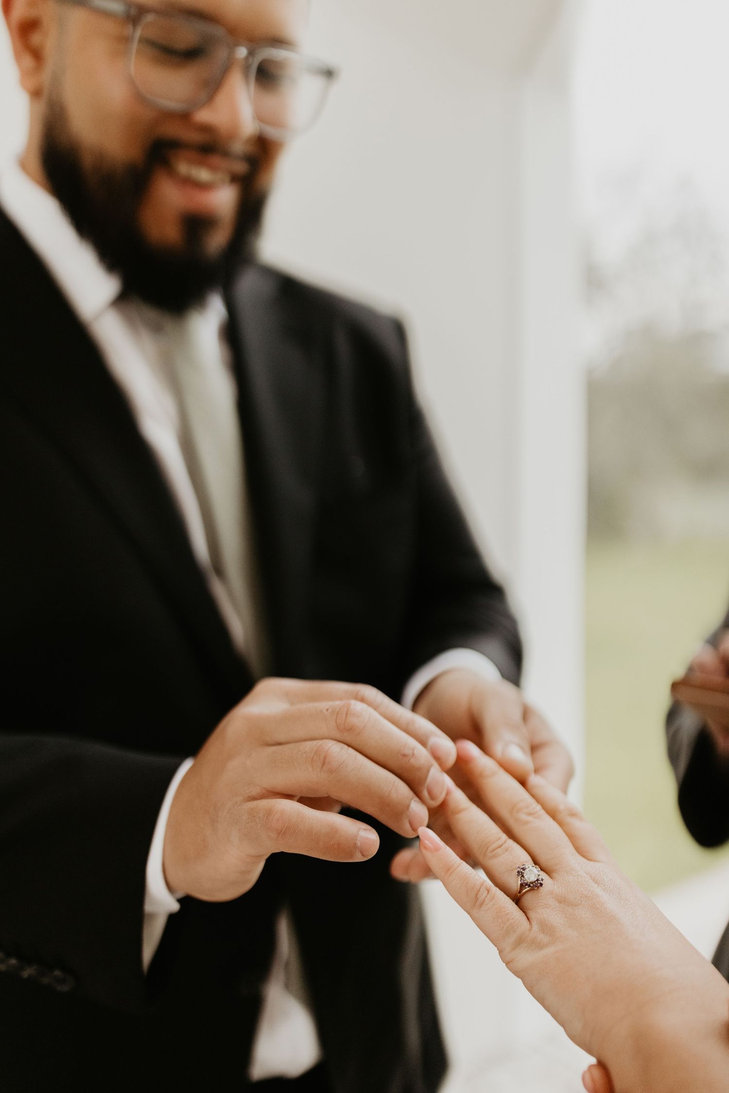 Close up of hands exchanging wedding rings during a marriage ceremony, with person wearing suit and glasses in background.
