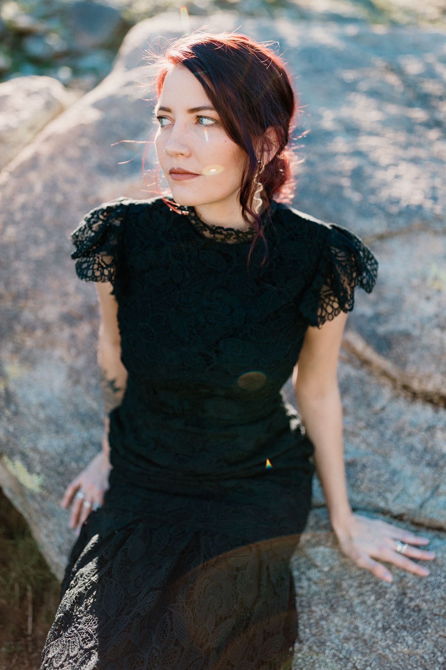 A woman in a black lace dress poses gracefully on rocky terrain during an outdoor photoshoot at sunset.