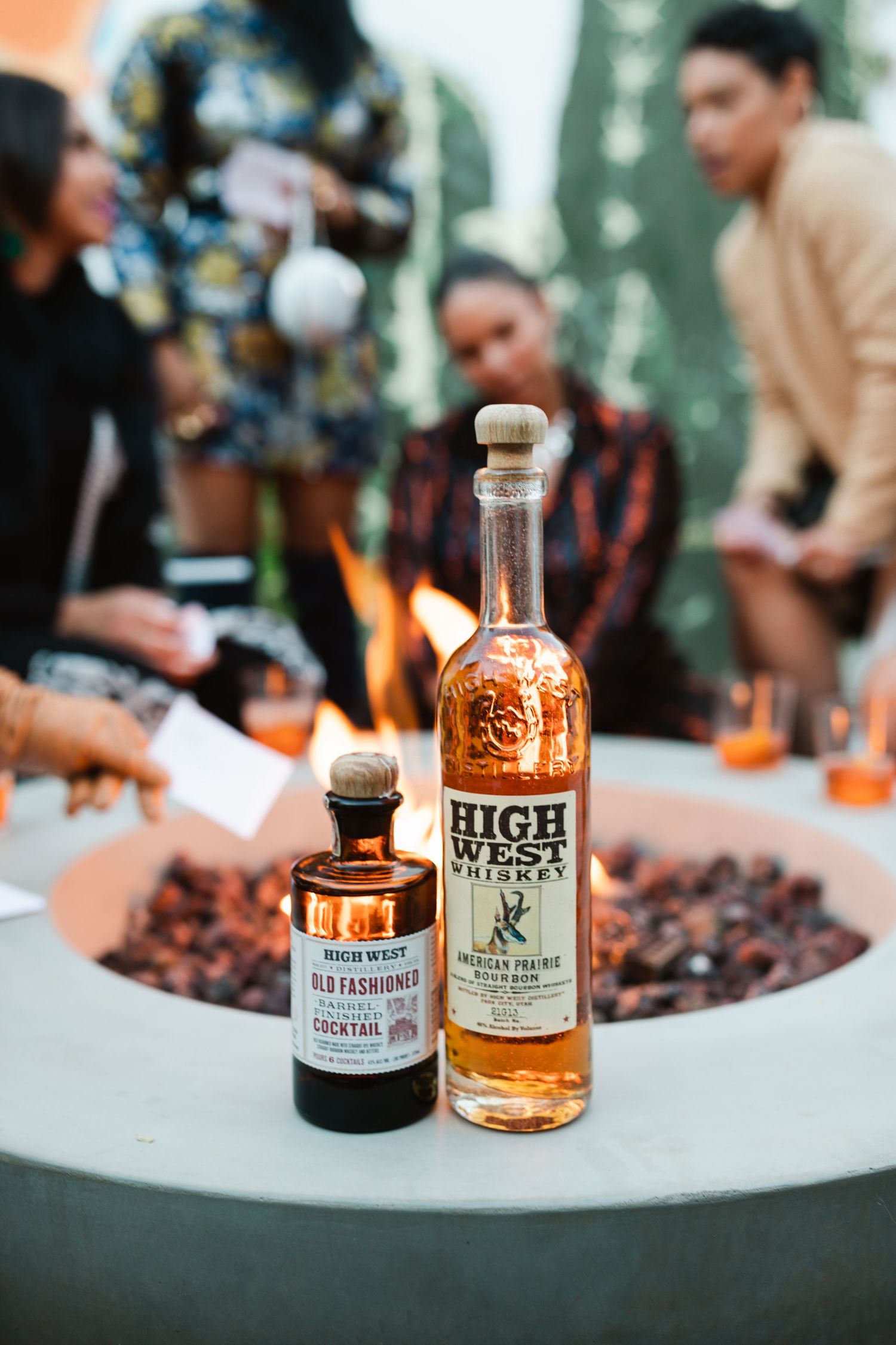 Two bottles of spirits sit on a white table with a blurred background of people gathering at an outdoor event.