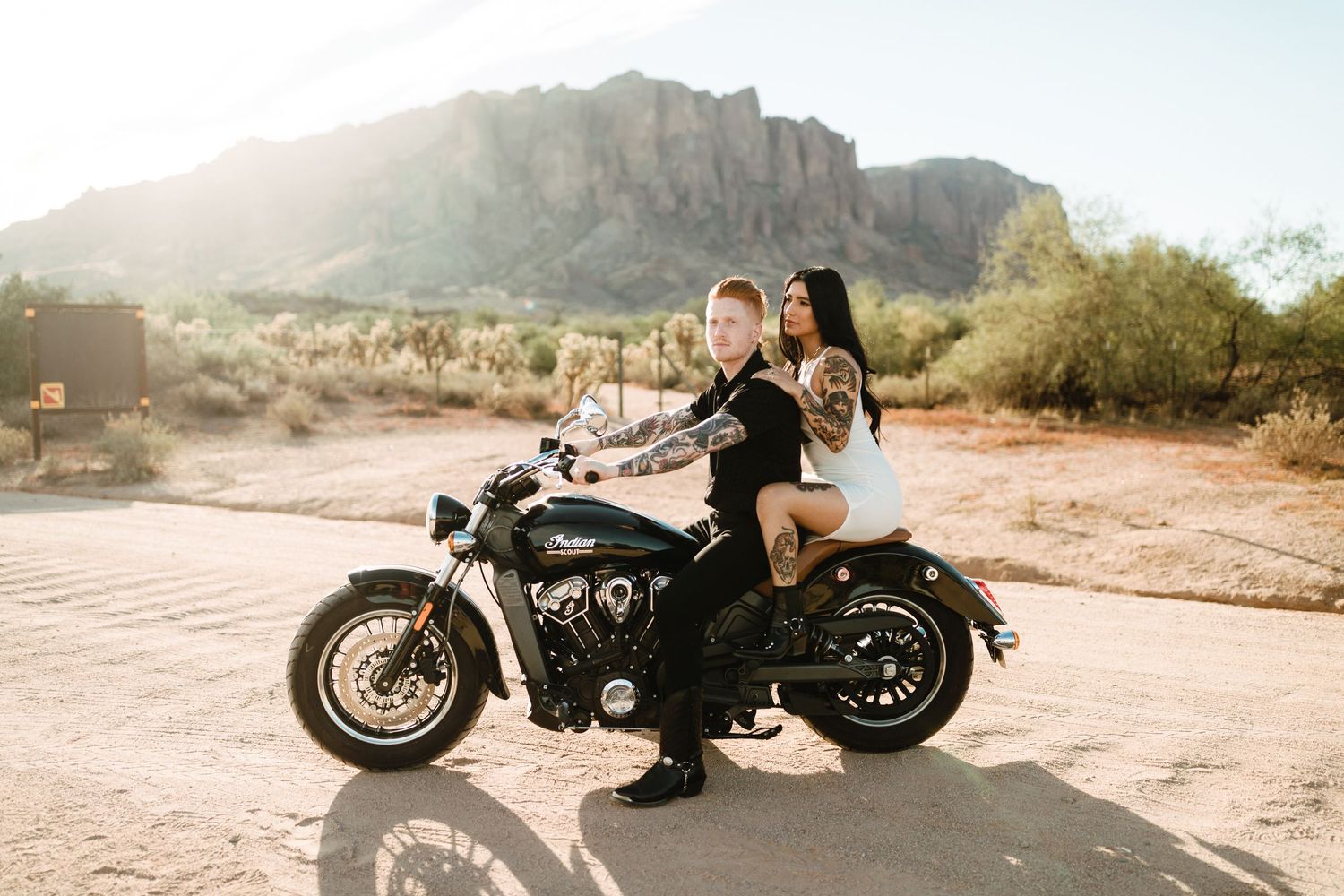 Motorcycle riders pose on bike in desert landscape with dramatic Superstition Mountains backdrop during golden hour.