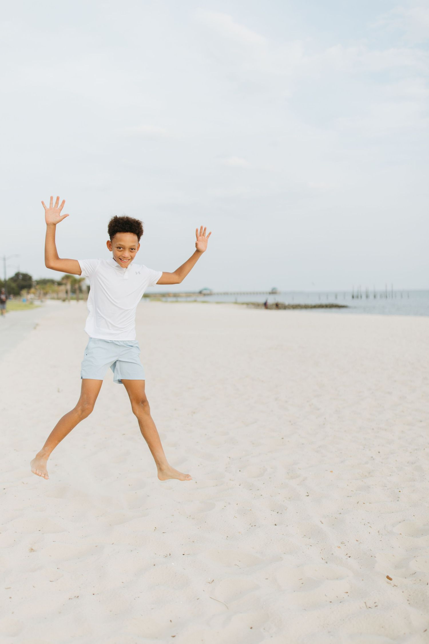 Young person in white shirt jumps playfully on pristine white sand beach with clear blue sky.