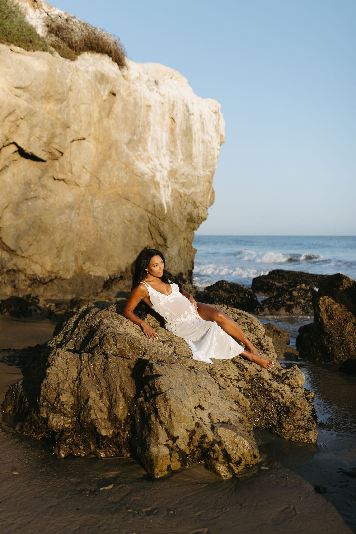 Person in a white dress lounges on coastal rocks at a beach with white cliffs and crashing waves in the background.