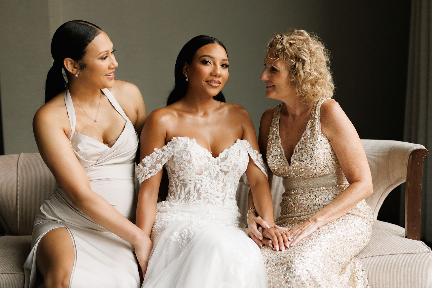 Three friends share a joyful moment on a couch wearing elegant formal gowns in white and sparkly tones.