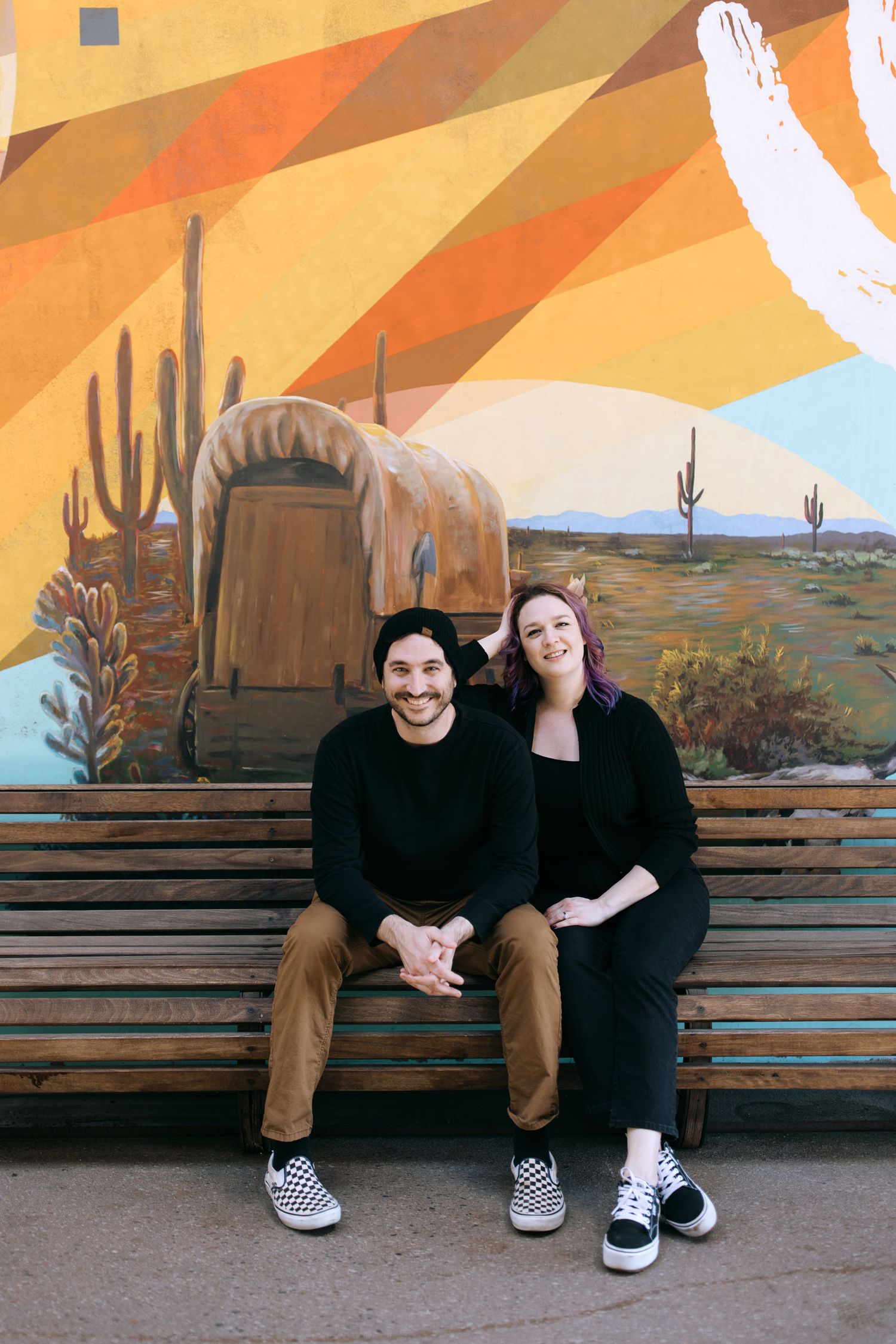 Two people sit together on a wooden bench with southwestern desert art and saguaro cactus murals painted on the wall behind them.