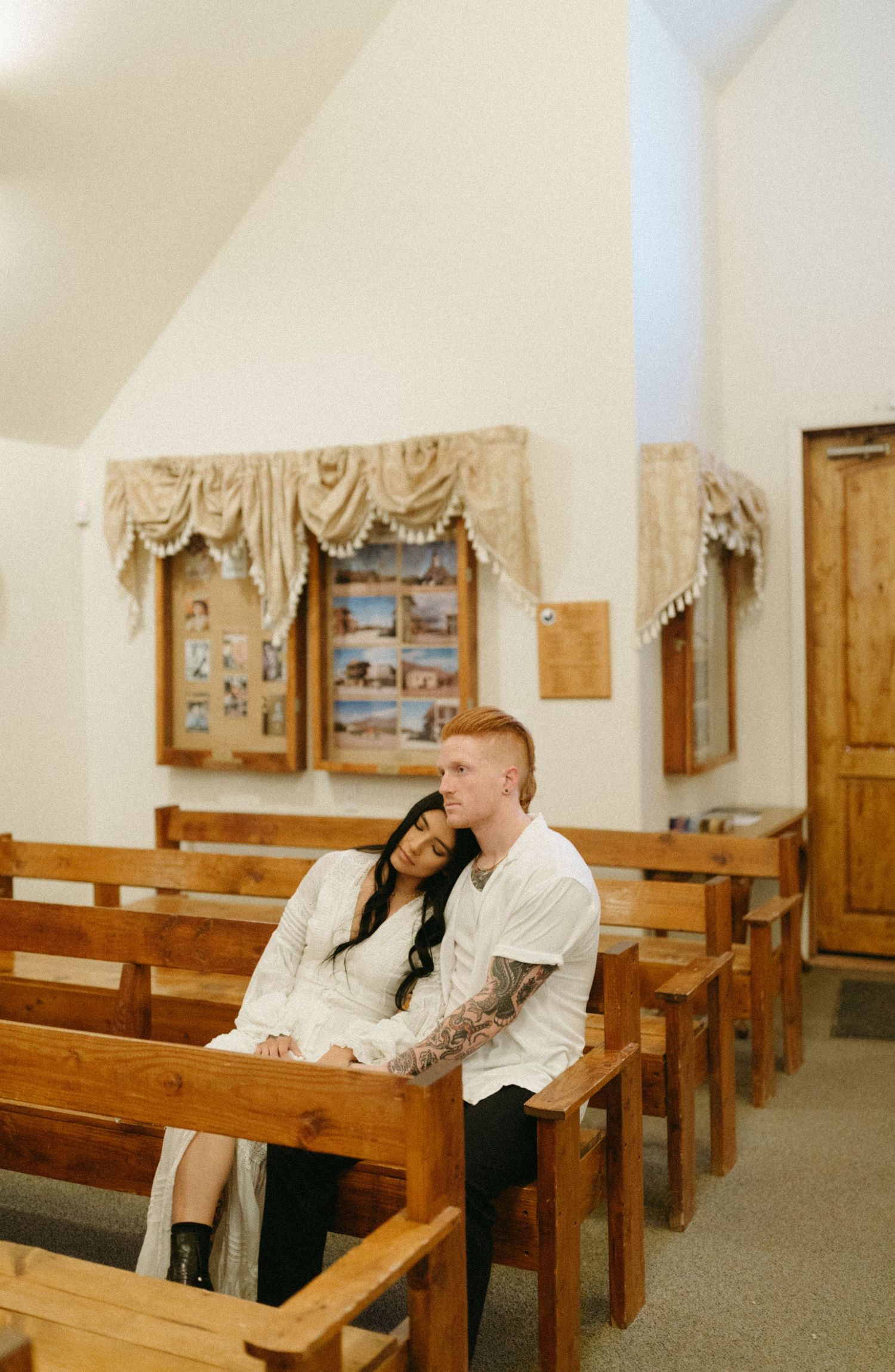 A couple shares an intimate moment while sitting together on wooden church pews in a simple chapel interior.