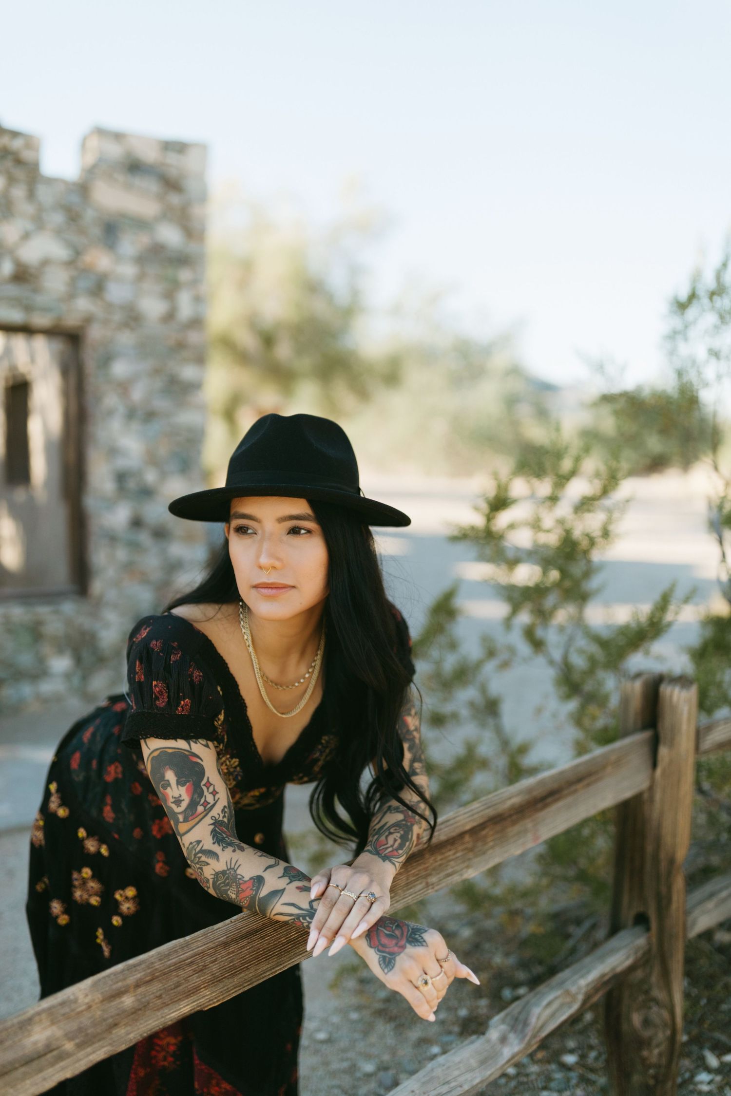Person in black lace dress and wide brim hat poses against rustic wooden fence in sunlit outdoor setting.
