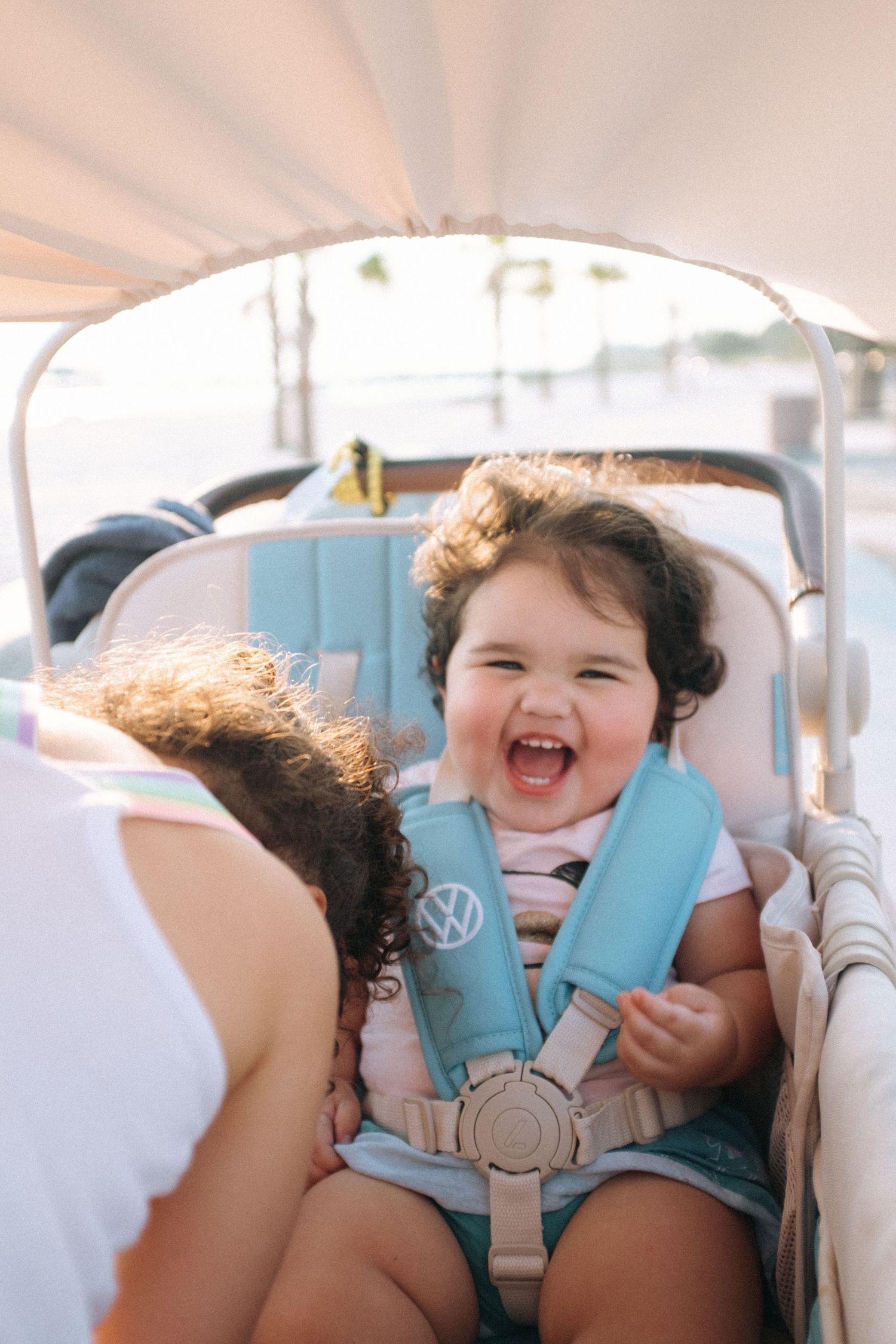 A laughing baby wearing a light blue safety harness sits strapped into a car seat.