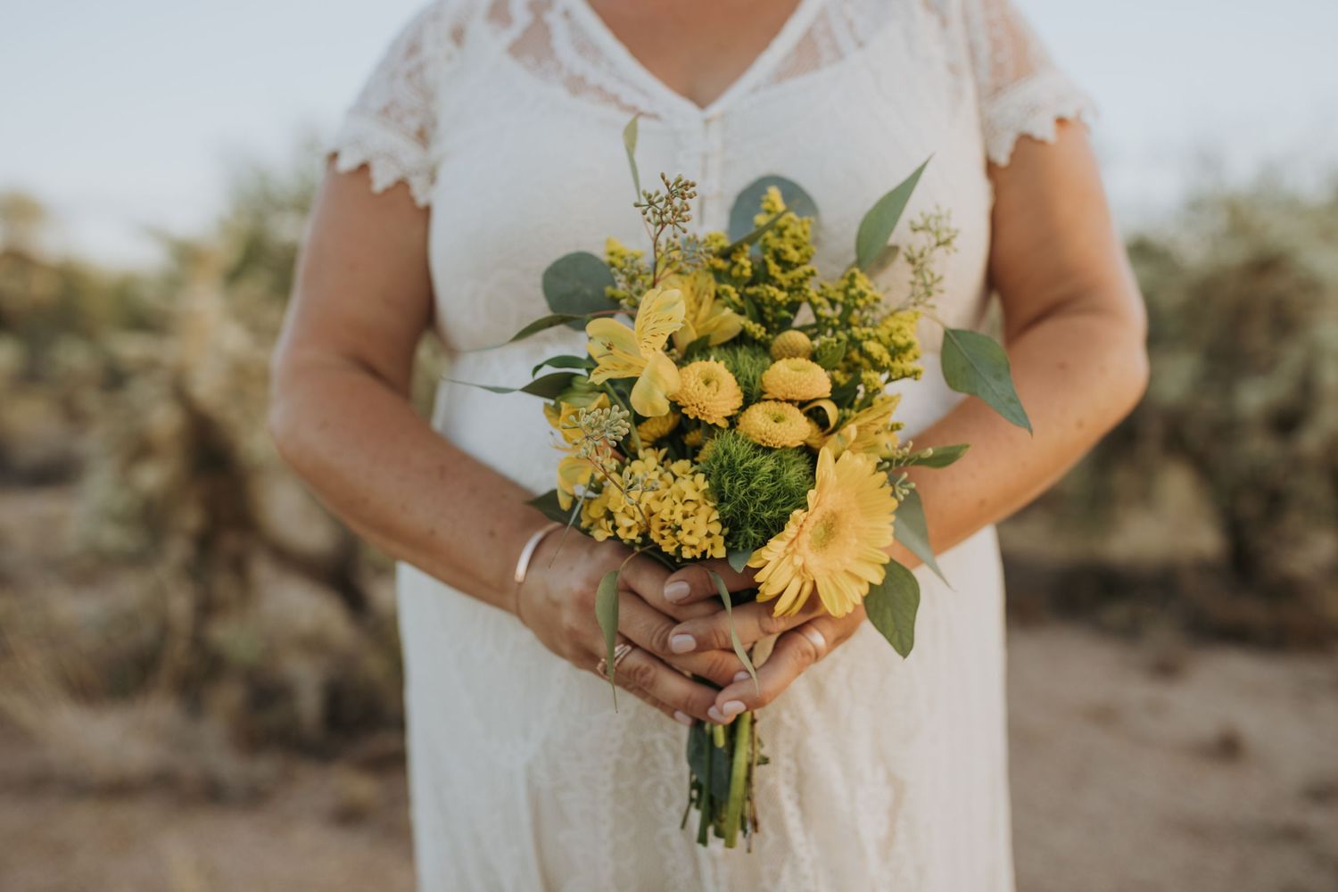 A wedding bouquet featuring yellow sunflowers, delicate white flowers, and green foliage held against a white dress.