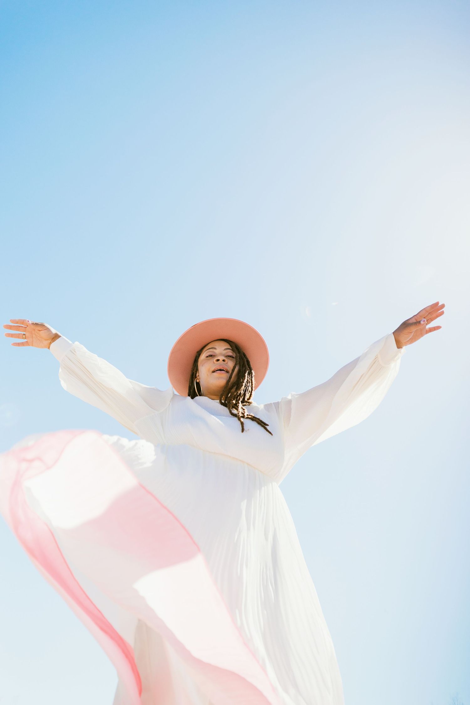 Person in white flowing dress and brown hat stands with arms outstretched against blue sky in joyful celebration pose.