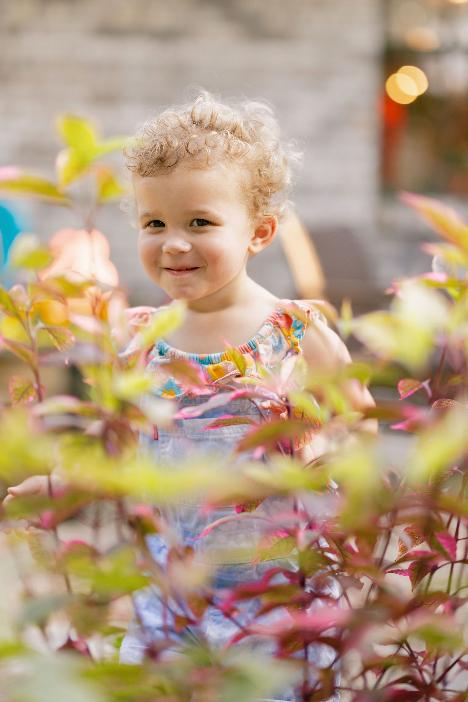 Young child peeking playfully through colorful spring foliage in a garden setting with soft bokeh background.