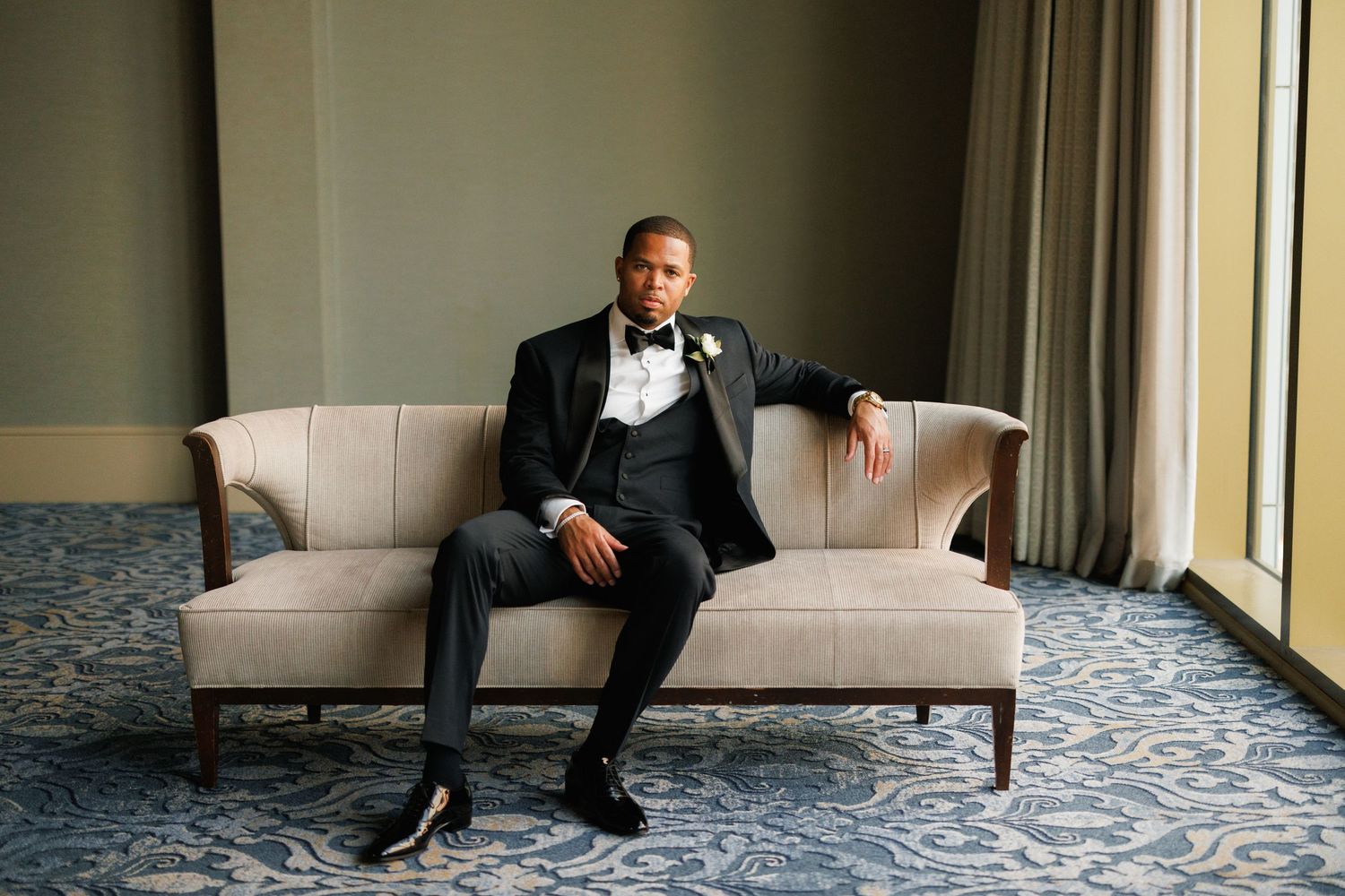 A person in a black suit sits on elegant beige sofa against a cream wall near large windows.