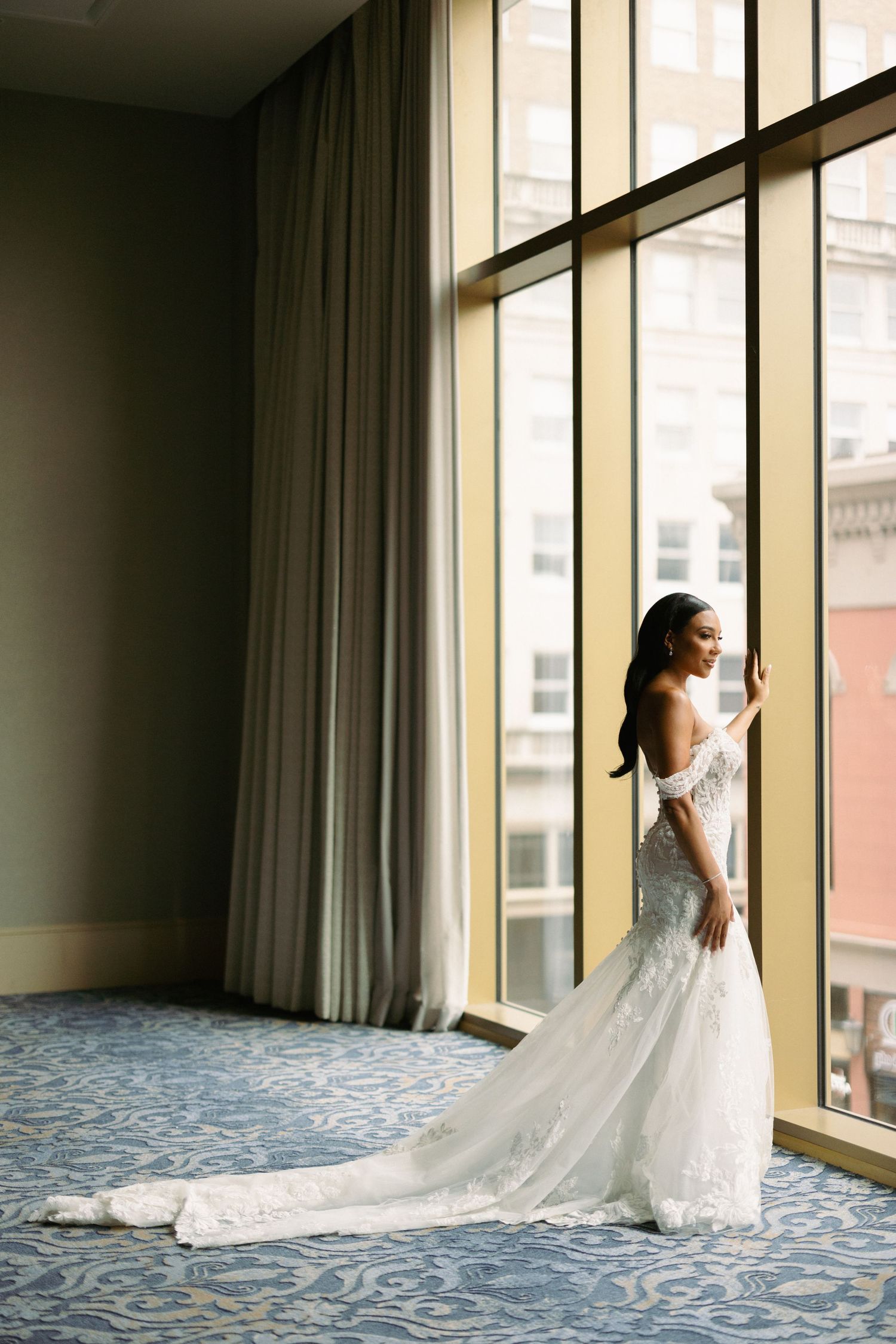 A stunning wedding gown with a flowing train is silhouetted against large windows in an elegant hotel room.