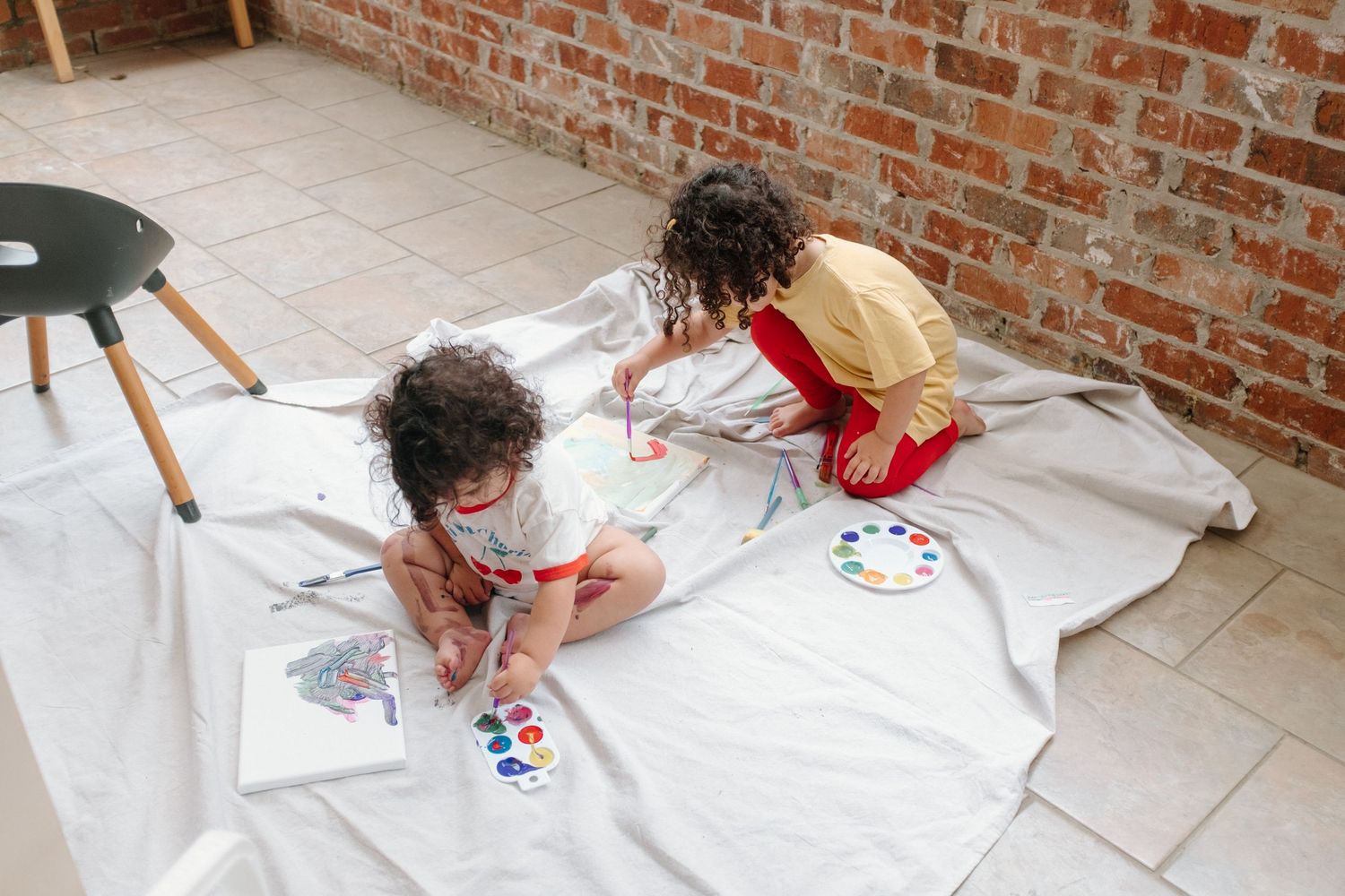 Small children lie on a sheet of white paper painting with red and blue paint on a brick patio floor.