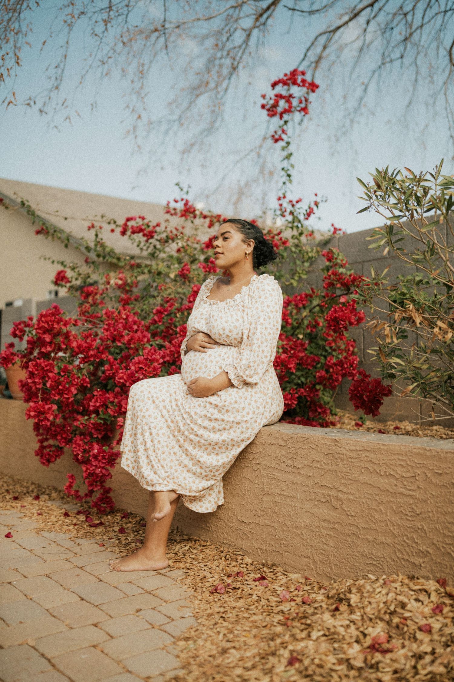 A person in a polka dot dress sits on a wall surrounded by vibrant red bougainvillea flowers in a garden setting.