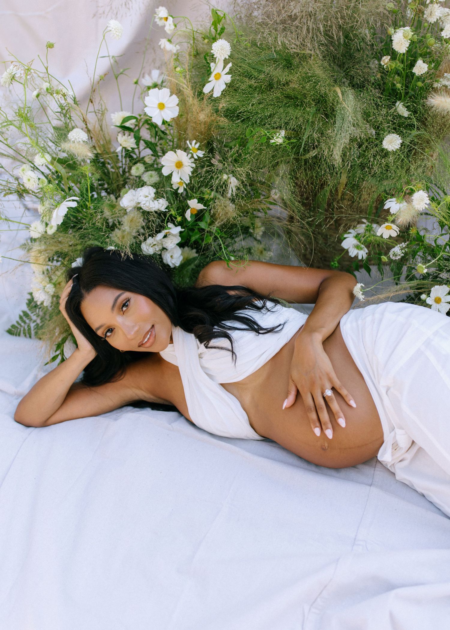 A person in a white dress lies gracefully among delicate white flowers and greenery in an artistic photoshoot setting.
