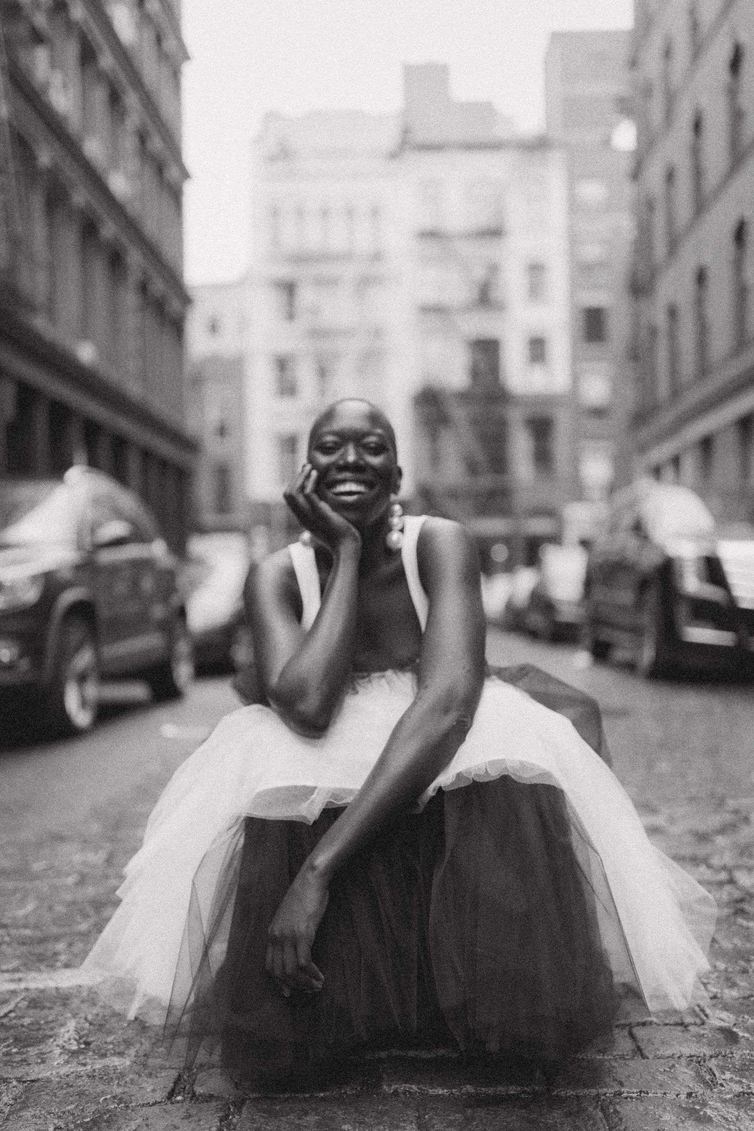 A ballerina in a white tulle dress sits joyfully on a city street with historic buildings and parked cars in the background.